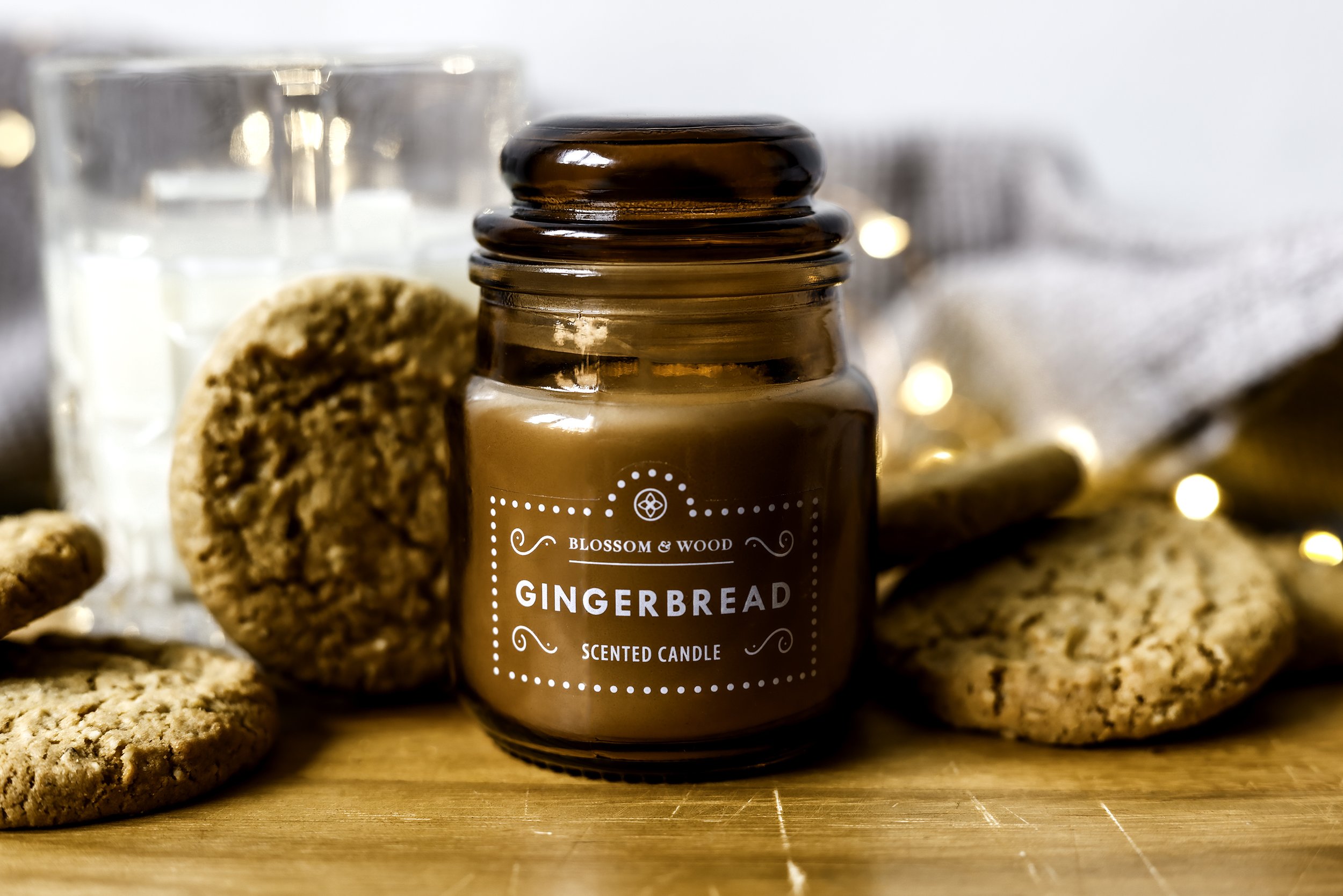 Gingerbread scented candle in brown jar surrounded by cookies and decorative lighting on a wooden surface.