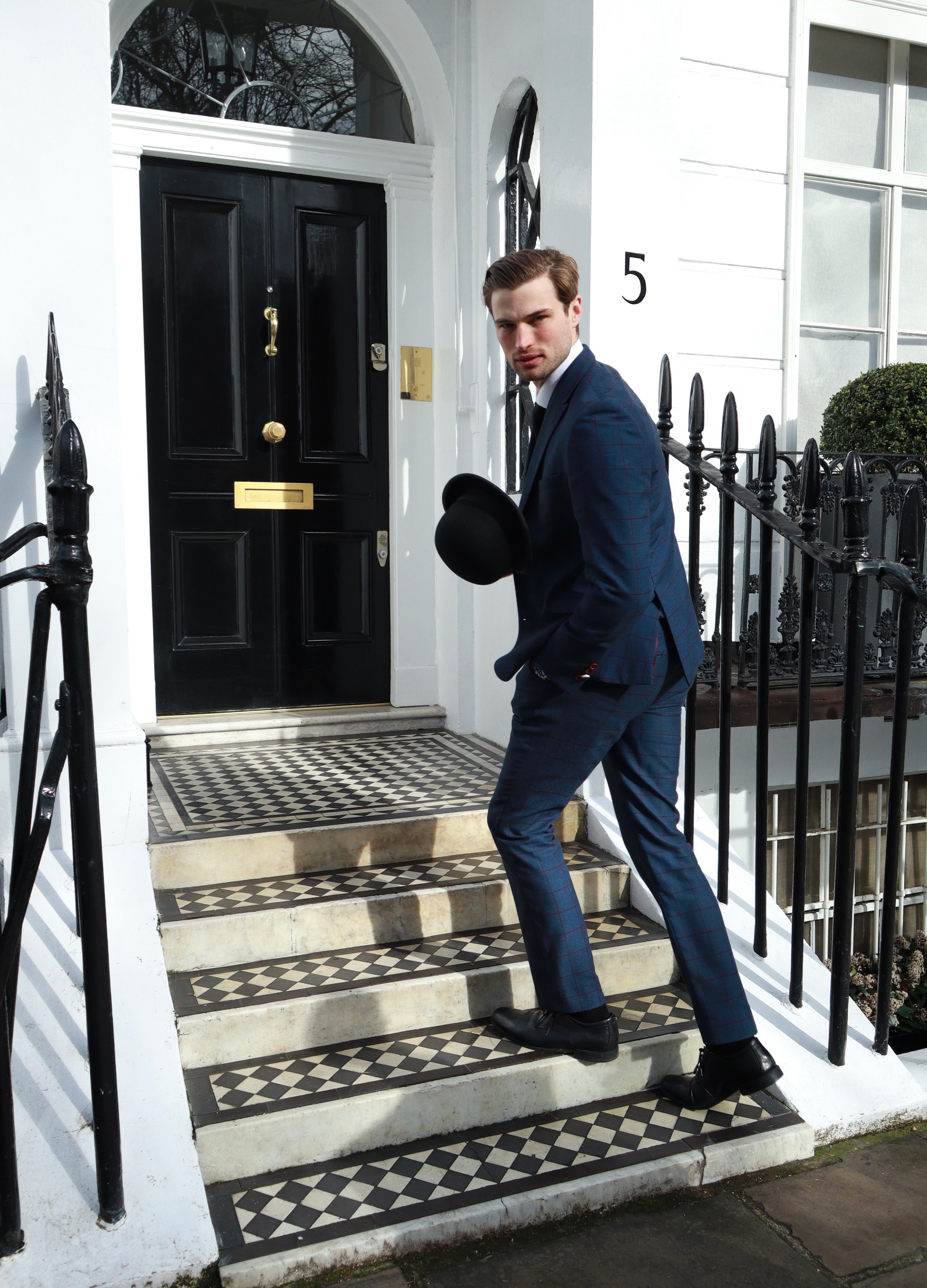 Man in blue suit holding black hat, walking up stairs in front of a white building with a black door and checkered steps.