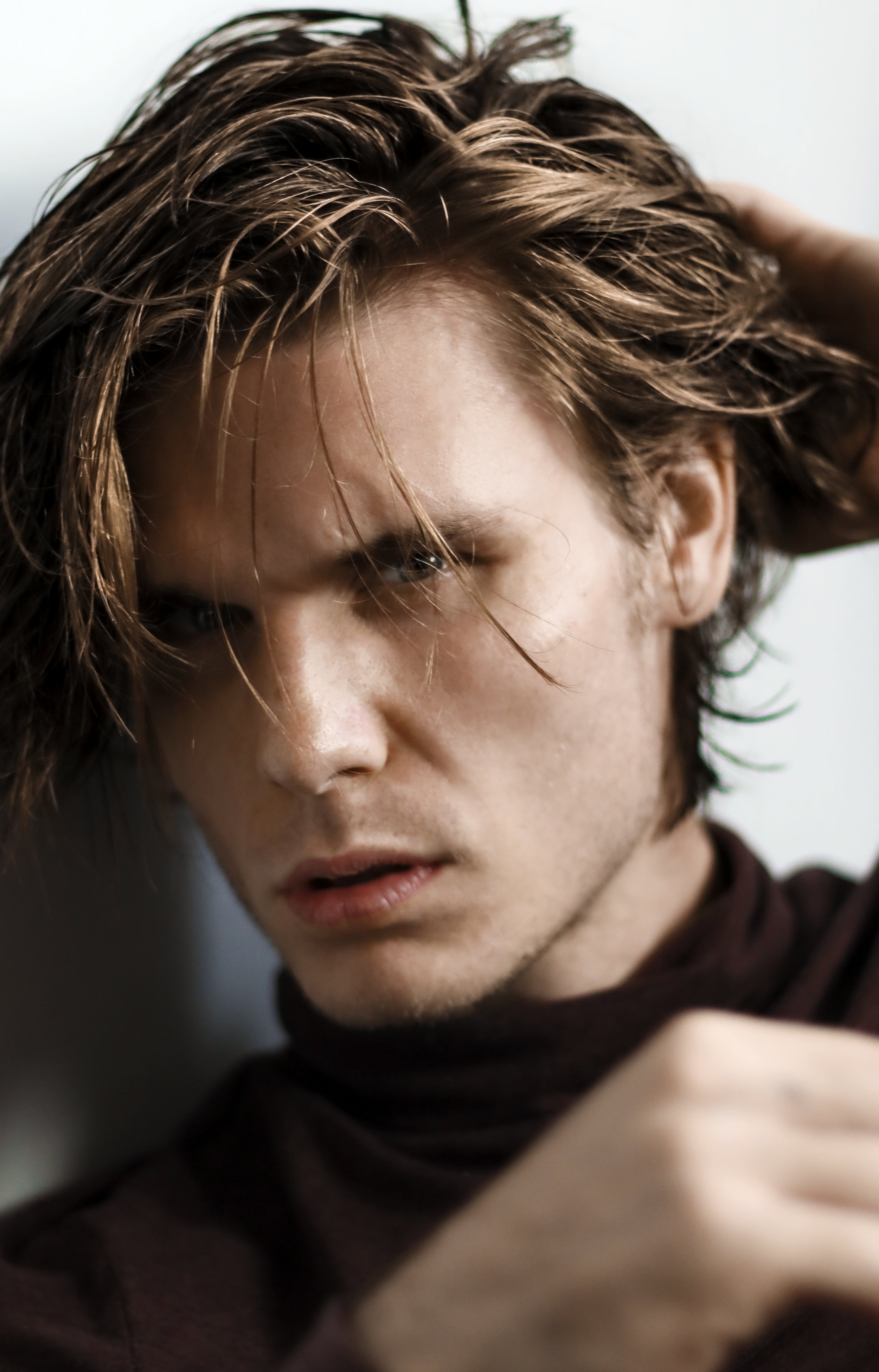 Close-up of a person with tousled brown hair, wearing a dark turtleneck, looking intently at the camera.