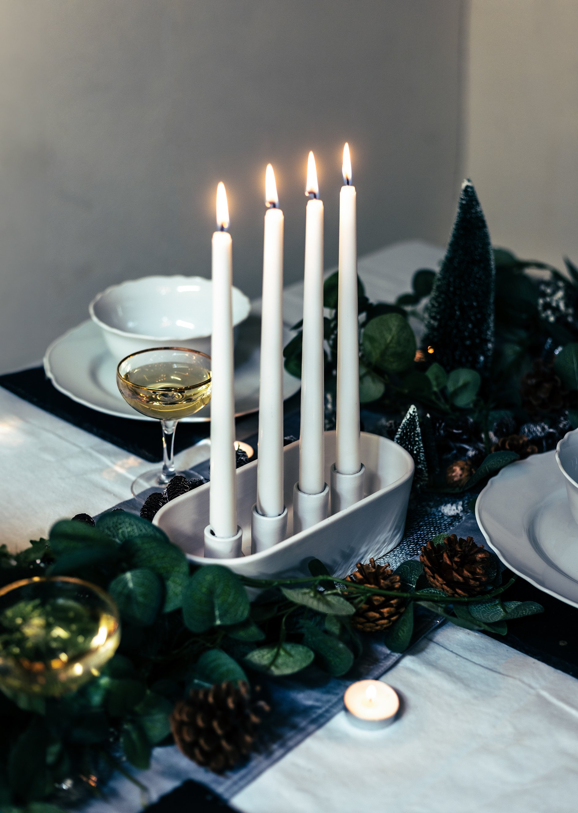 Elegant dinner table setup with four lit taper candles in a holder, surrounded by greenery and pinecones, plates, and glassware with a drink.