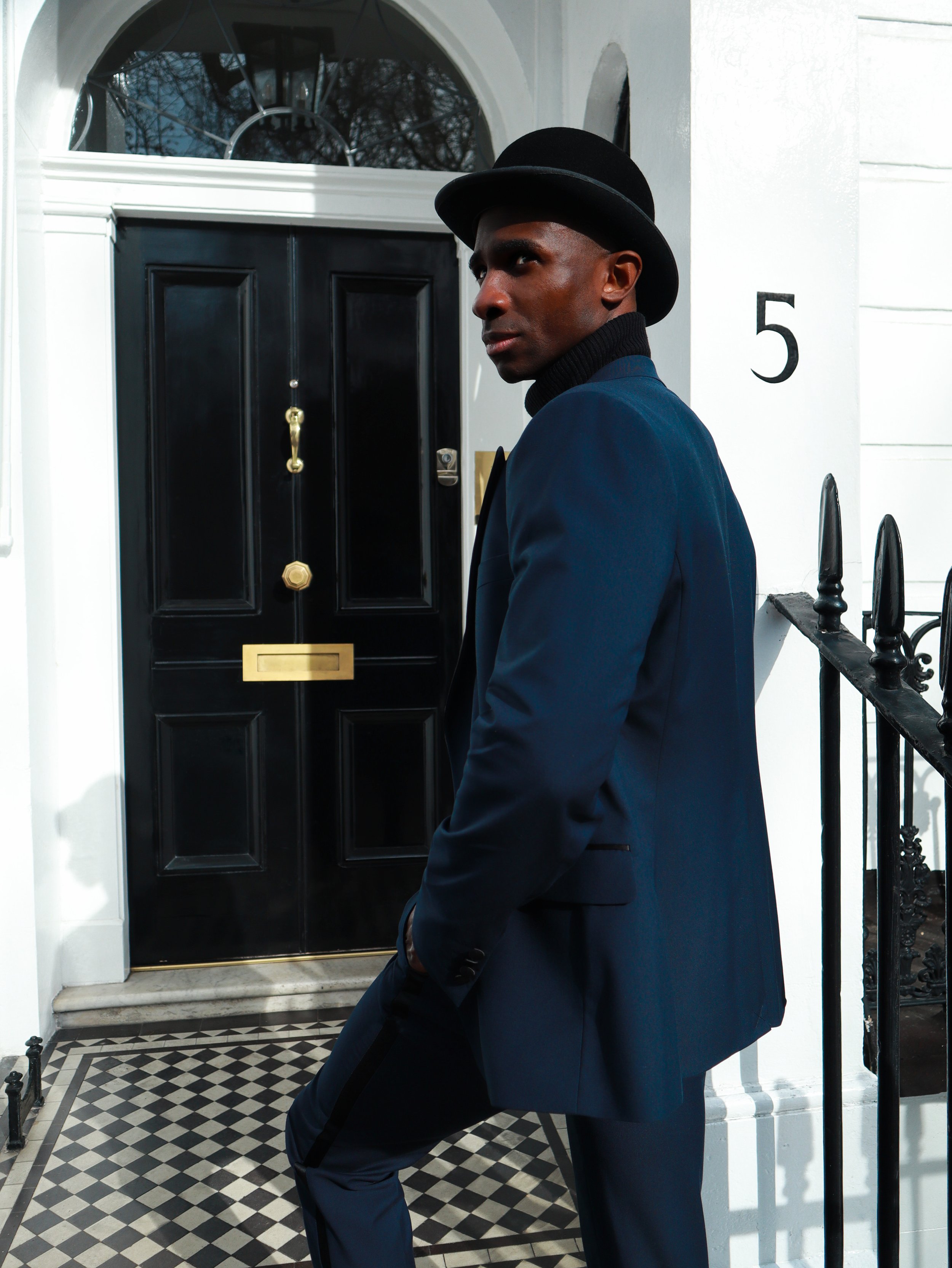 Man in a dark suit and bowler hat standing in front of a black door with number 5, on a checkered tile floor.