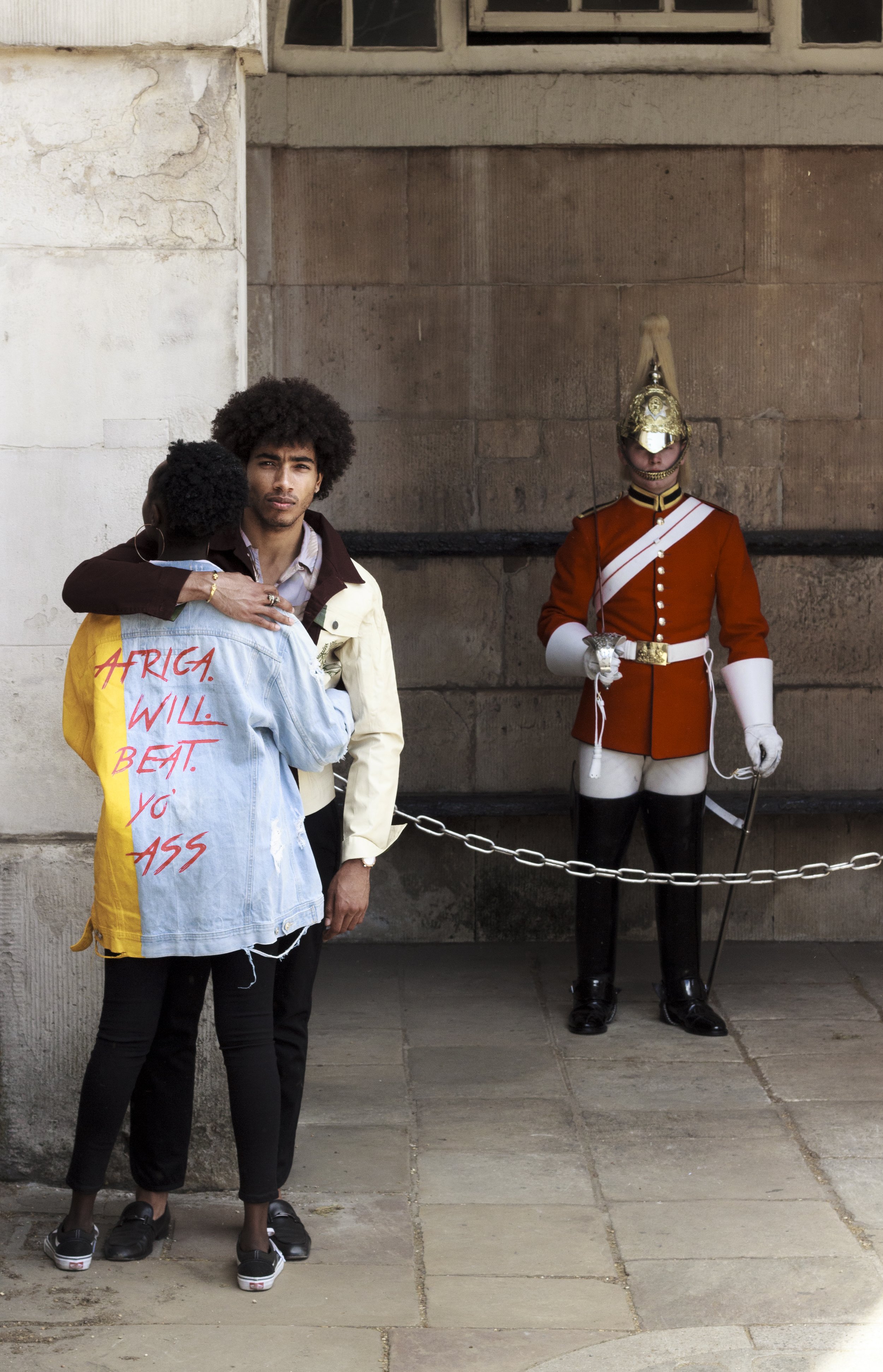 A person embracing another in front of a British guard in traditional uniform, with a message on one person's jacket.