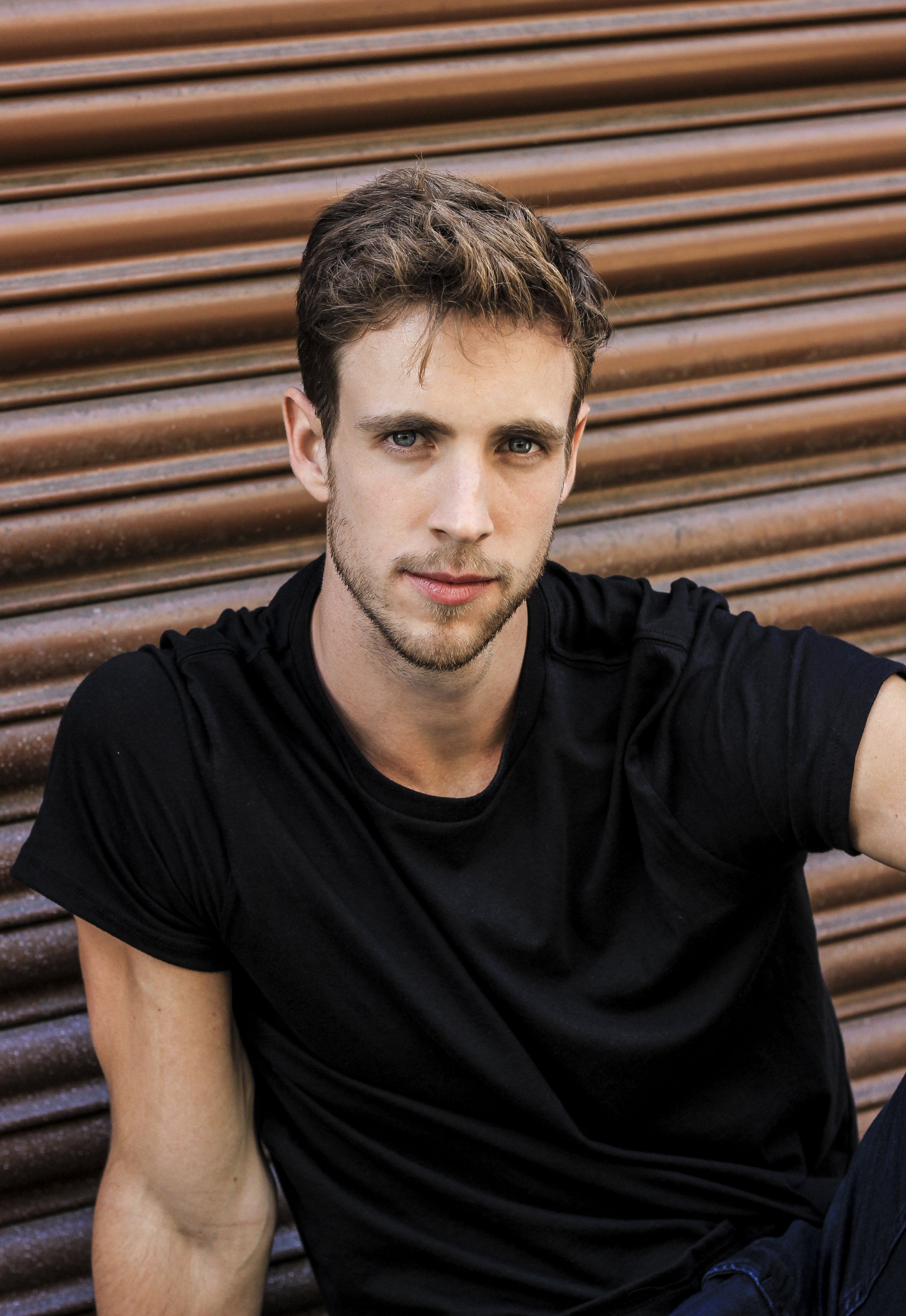 Man in black shirt sitting in front of a brown, corrugated metal background.