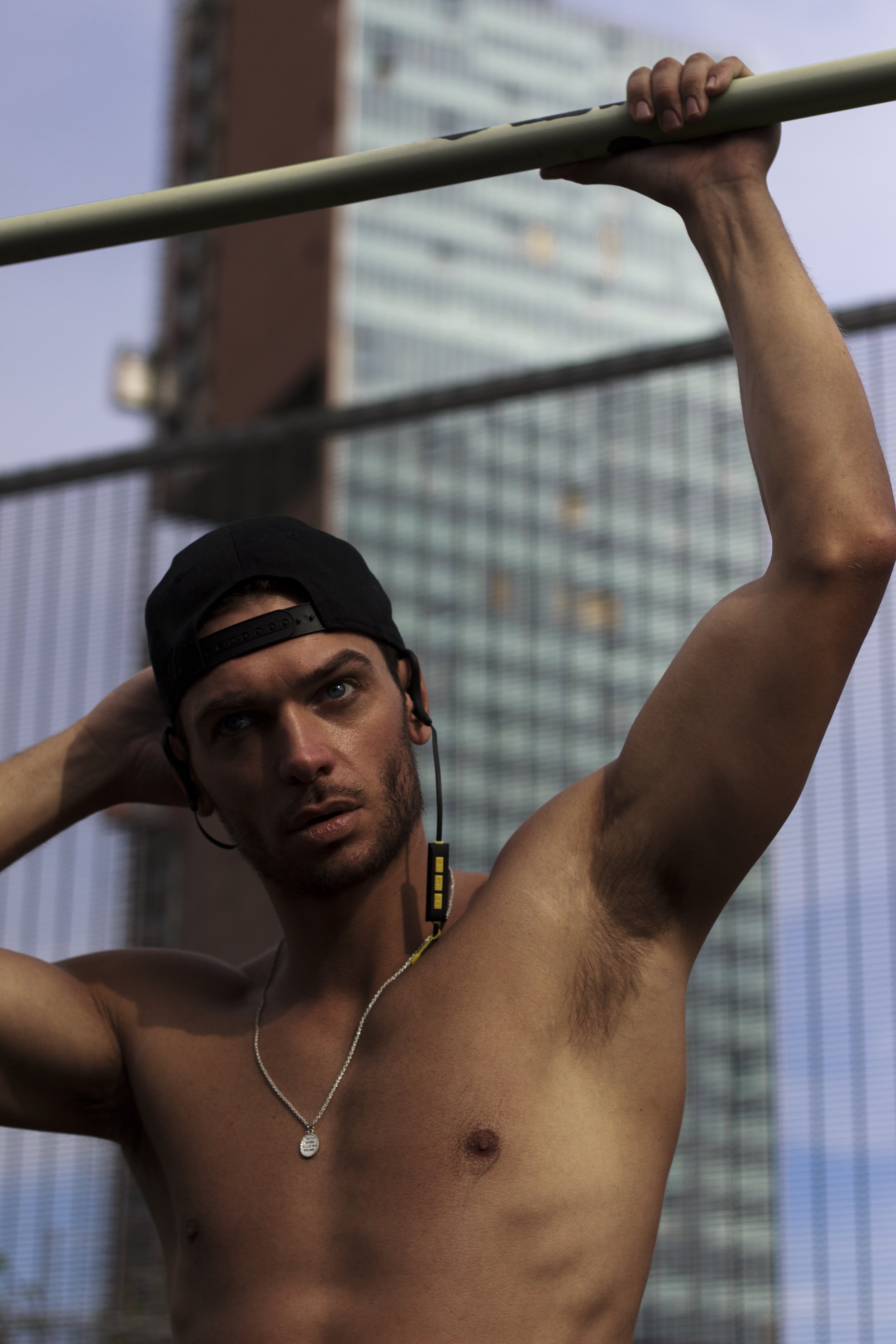 Shirtless man wearing a cap and necklace, holding a bar overhead, with earbuds and a cityscape in the background.