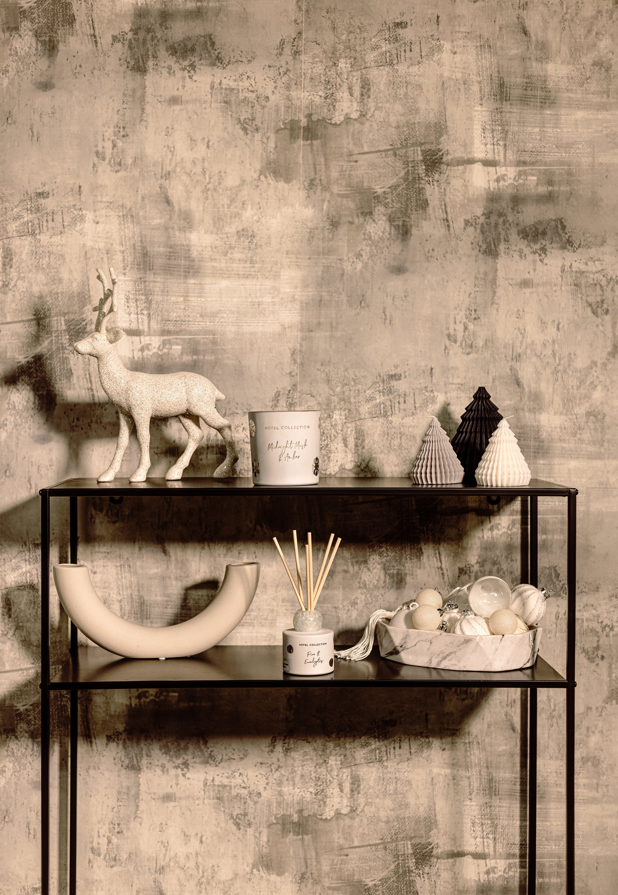 Decorative shelf with a reindeer figurine, candle, small tree sculptures, and a bowl with ornaments against a textured wall.