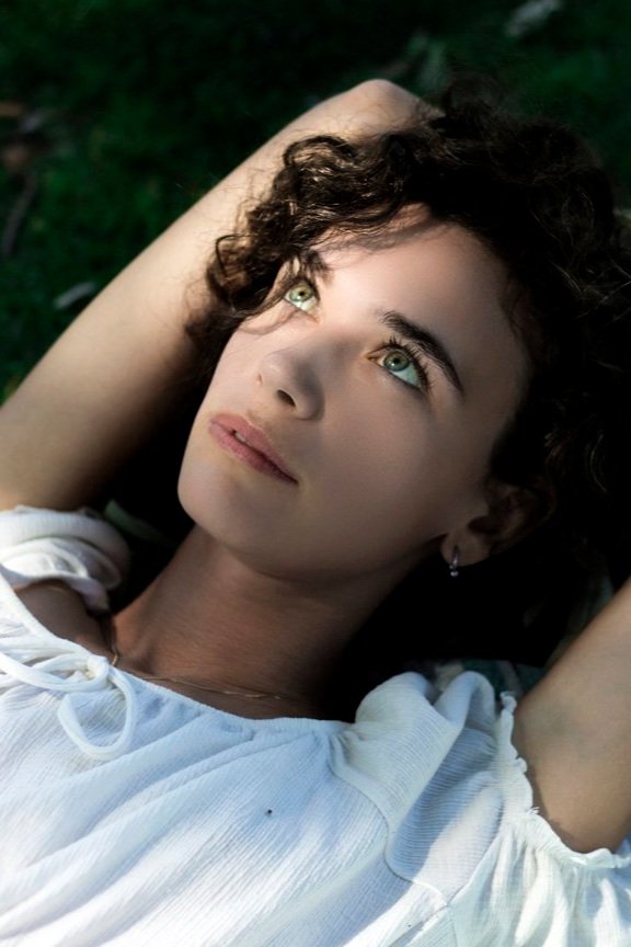 Person with curly hair lying on grass, gazing upwards, wearing a white blouse.
