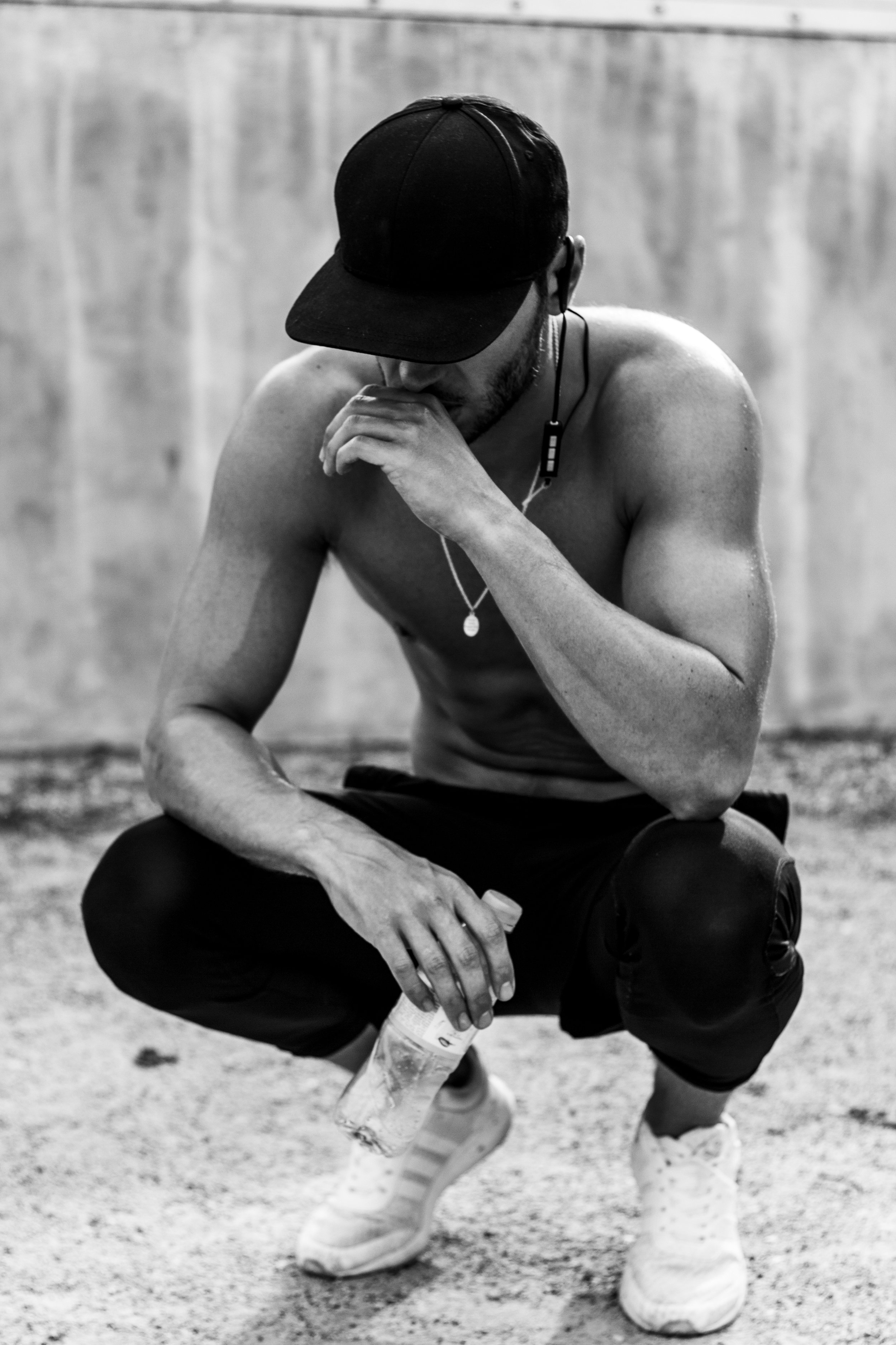 Black and white photo of a shirtless man squatting, wearing a cap, with headphones around his neck, holding a water bottle, and wearing sneakers.