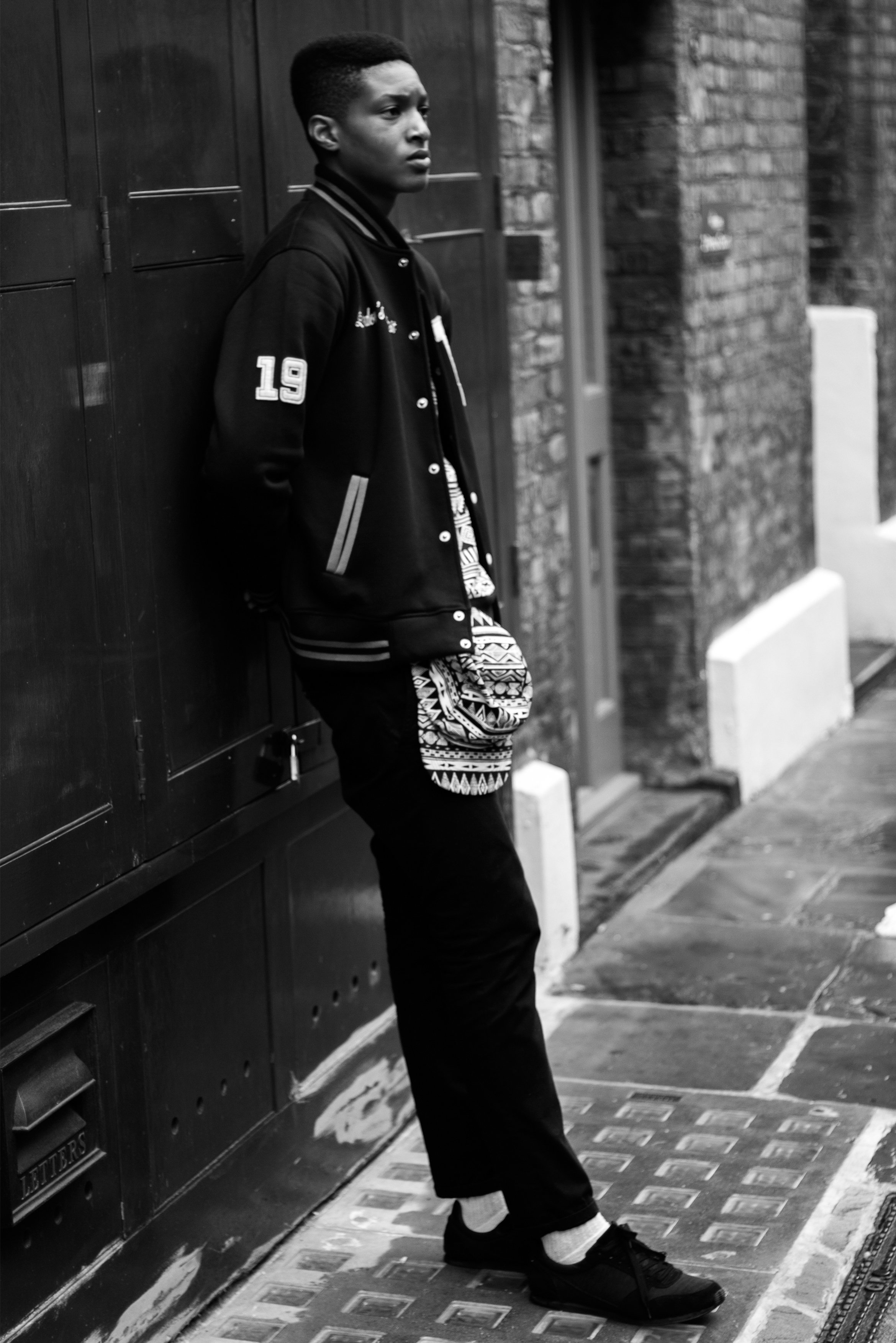 Black and white photo of a person leaning against a wall in an urban setting. They are wearing a letterman jacket with the number 19 on the sleeve, black pants, and sneakers. The person has a calm expression and is looking away from the camera.