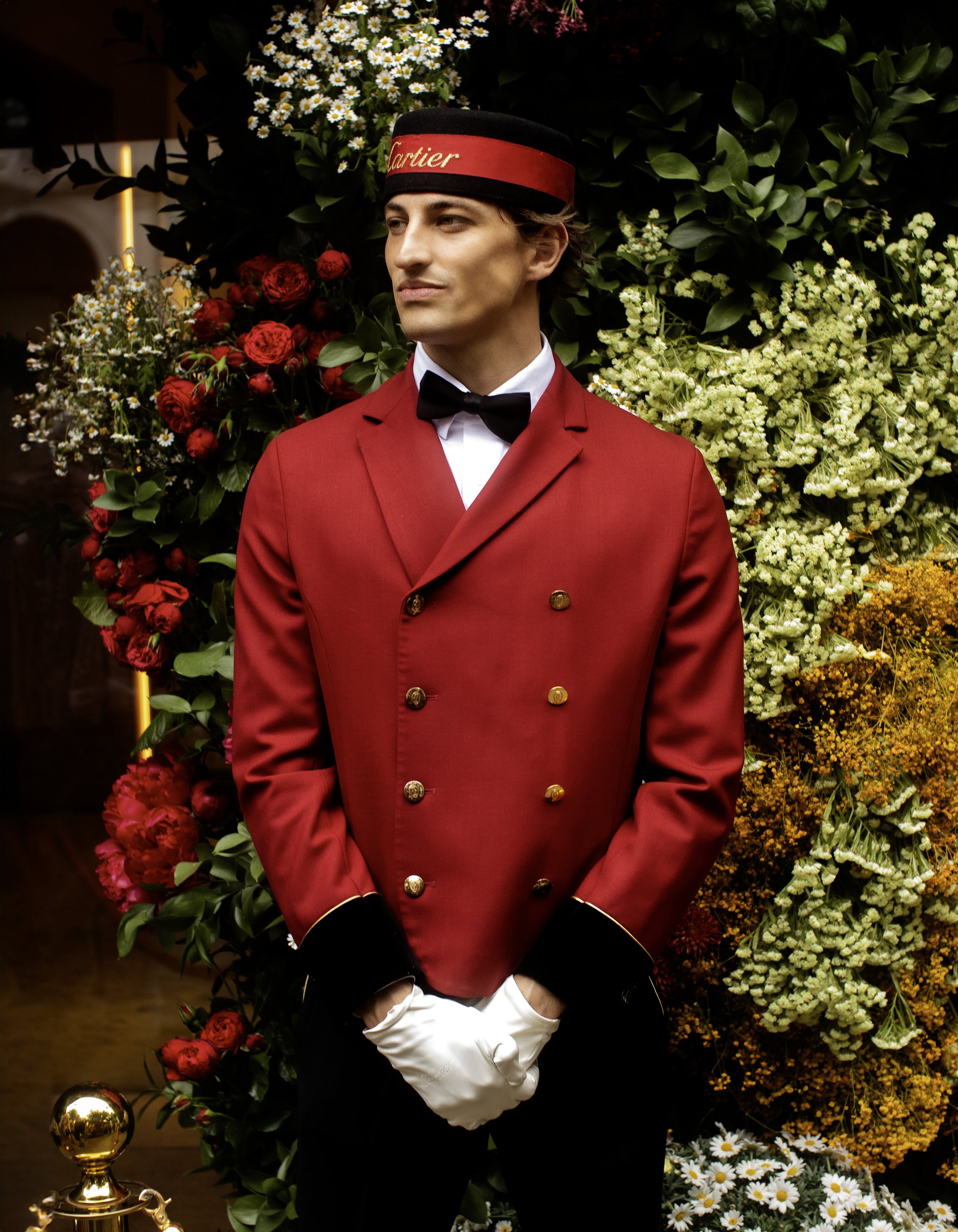 A doorman in a red uniform with a hat and gloves standing in front of a floral display.