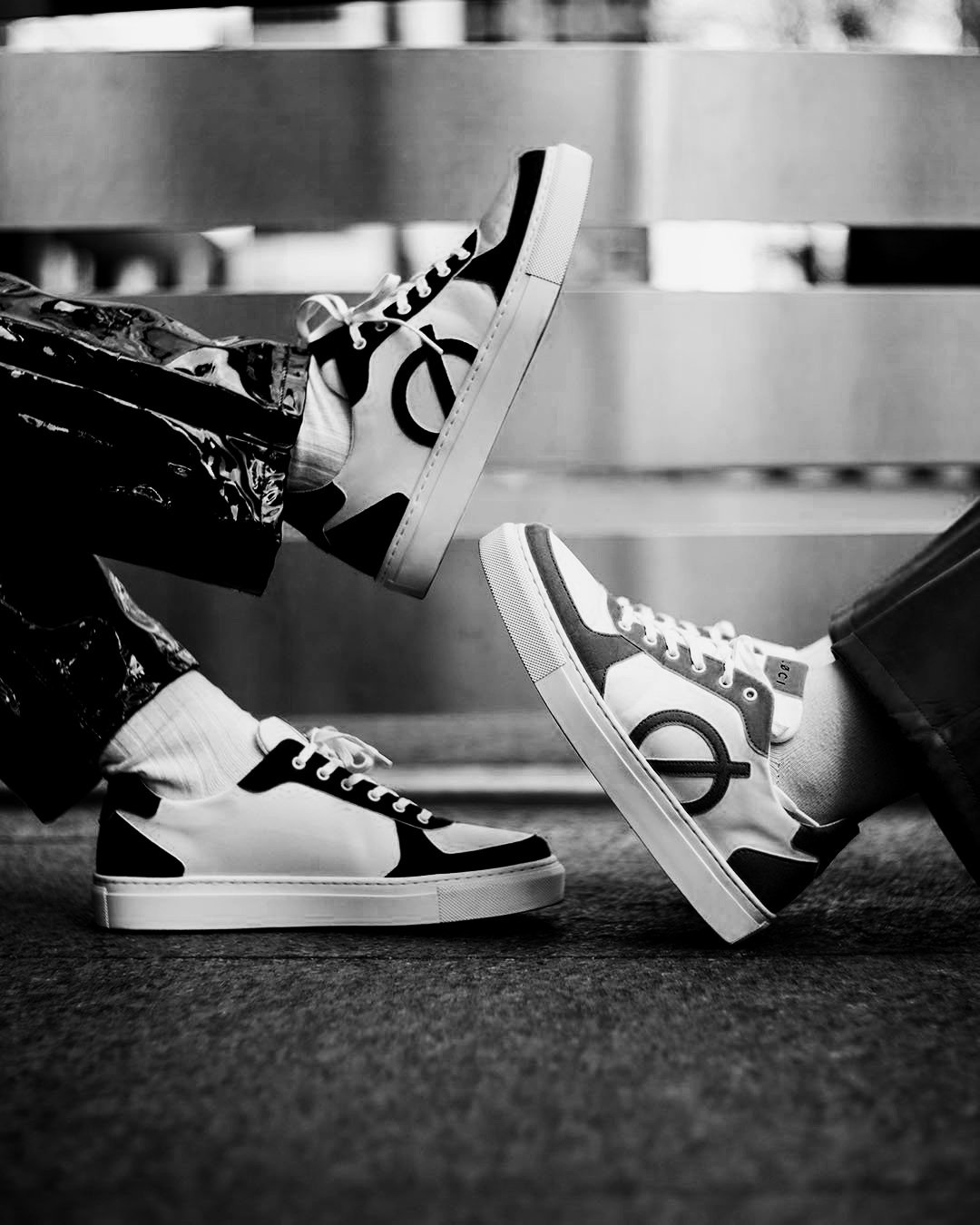 Black and white photo of two people wearing matching sneakers with a circular logo on the side, standing on textured ground, facing each other.