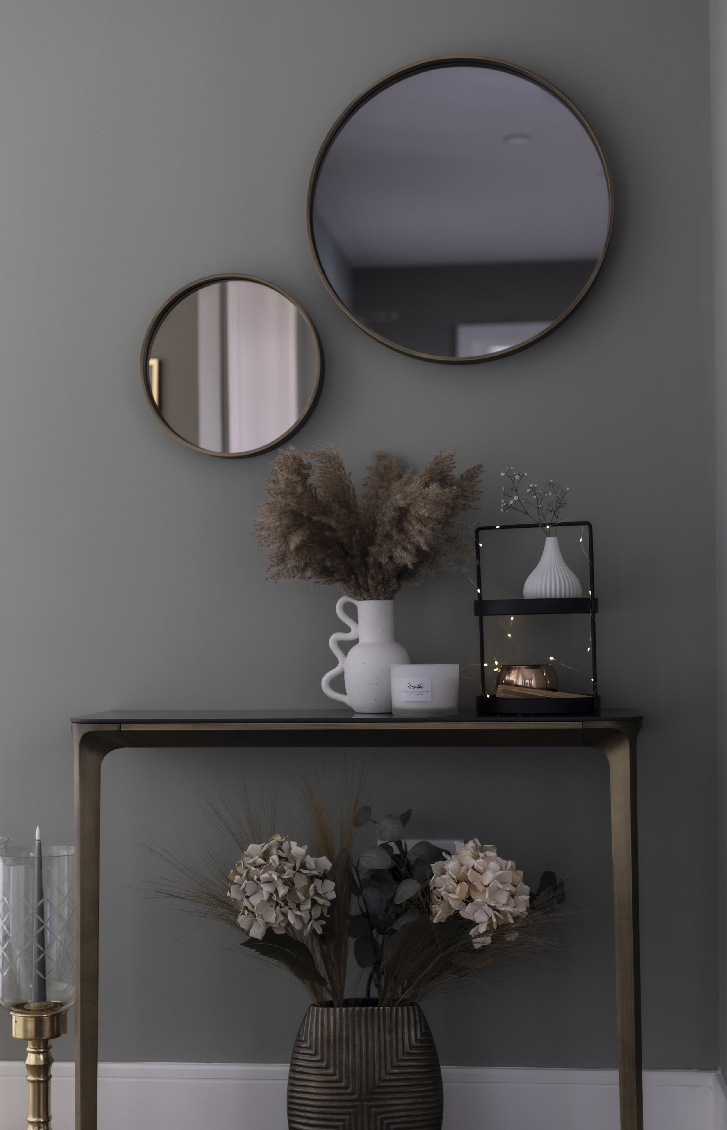 Interior decor with two wall mirrors above a console table, featuring a vase with pampas grass, a small decorative arrangement with lights, and floral decor in a textured vase below. Gray walls and modern style.