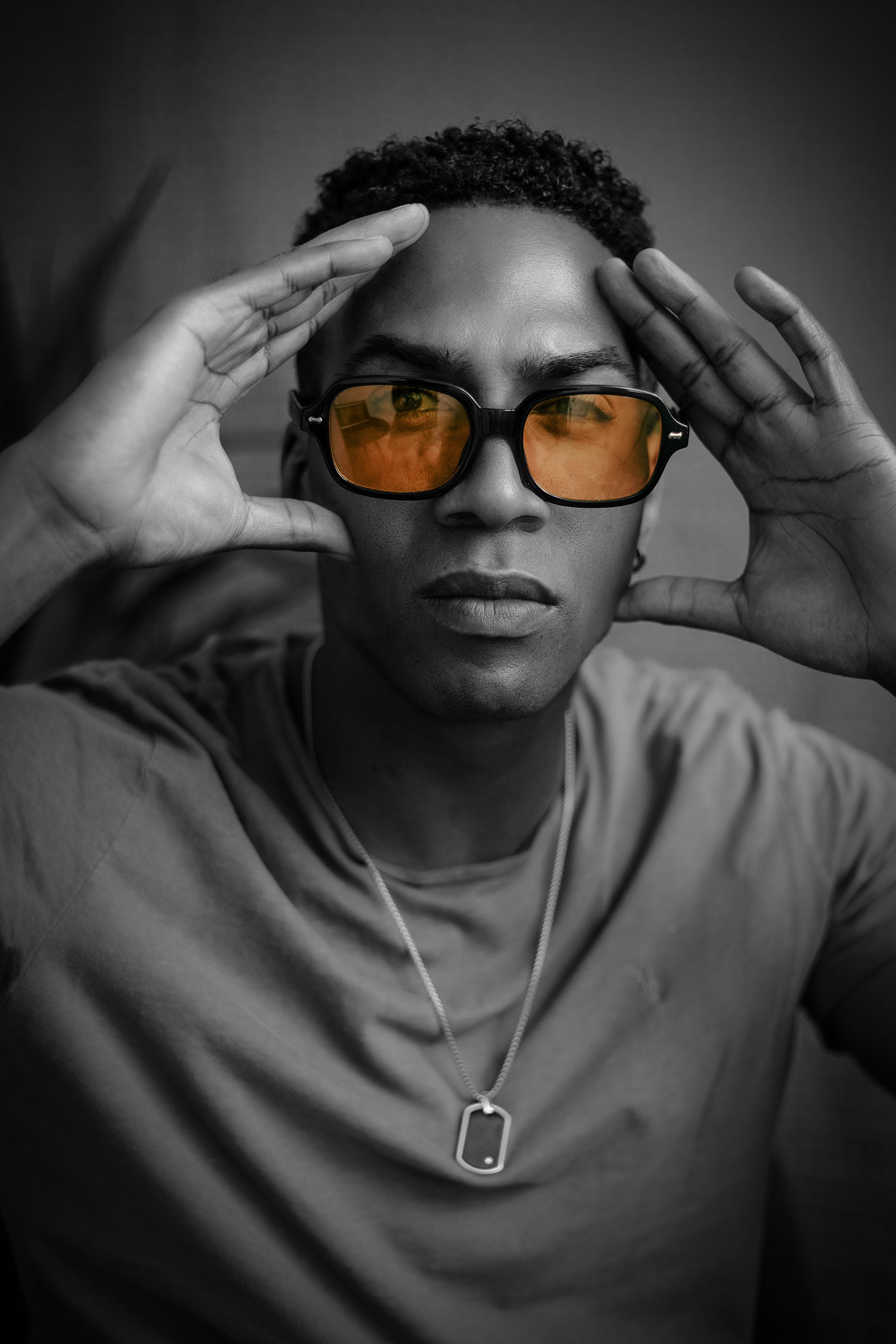 Man wearing orange-tinted sunglasses with hands framing his face, in a black and white setting.