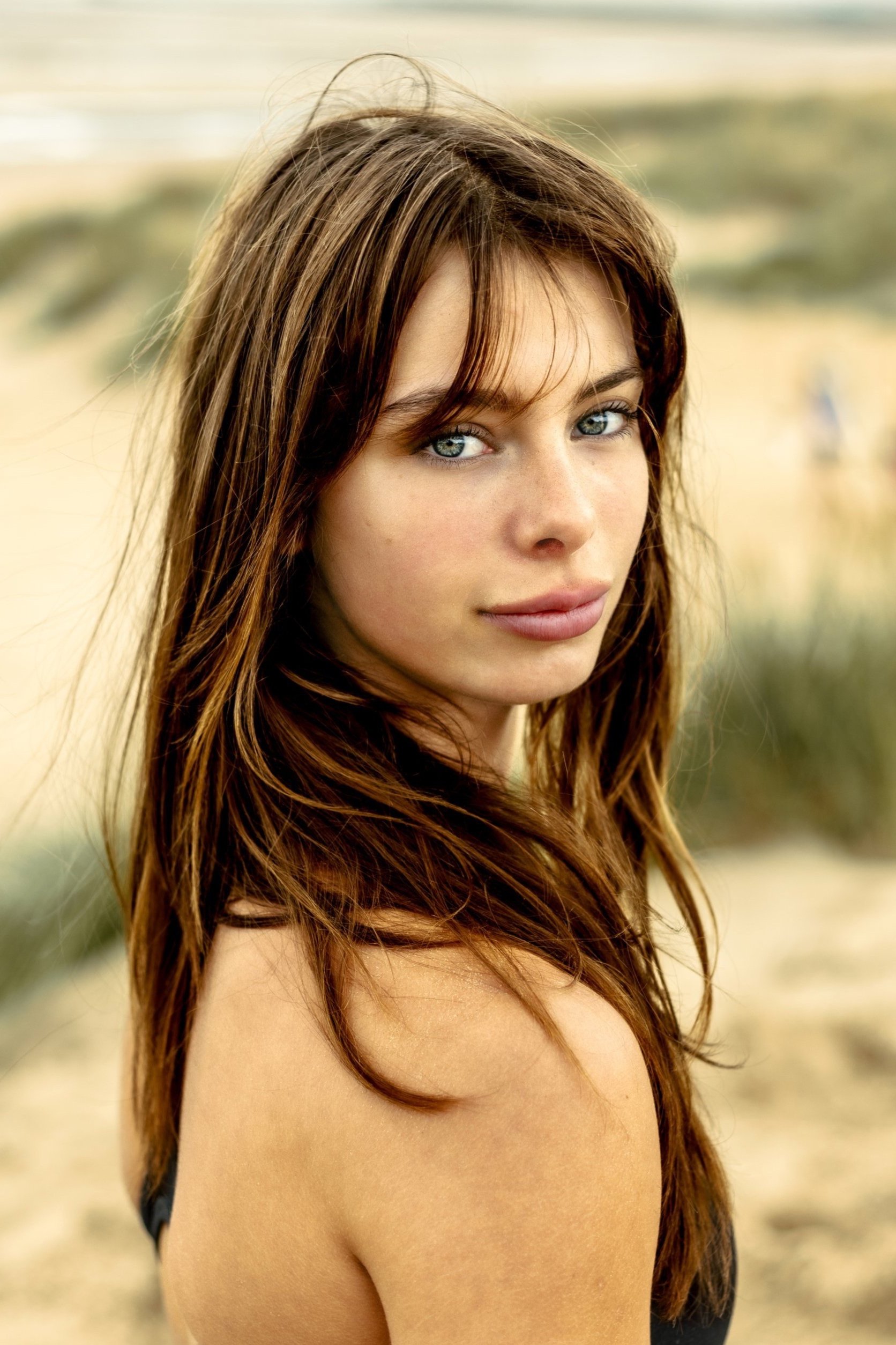 A woman with long brown hair and blue eyes looking over her shoulder at the camera, standing on a sandy beach.