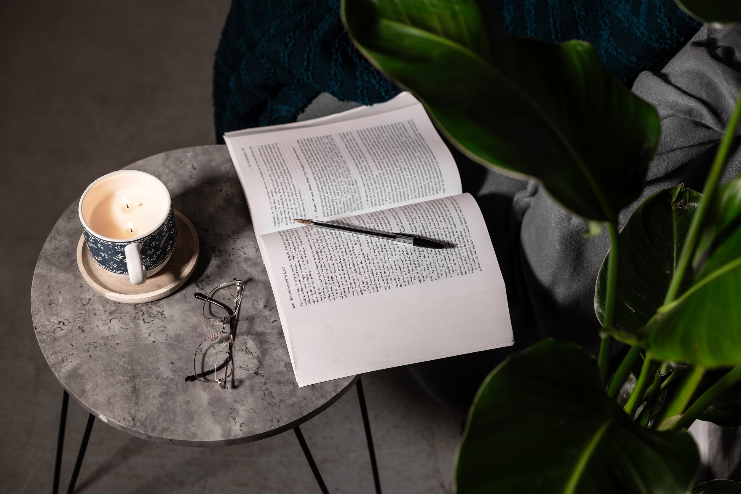 Open book with a pen on it, glasses, candle on a small table, and green plant leaves.