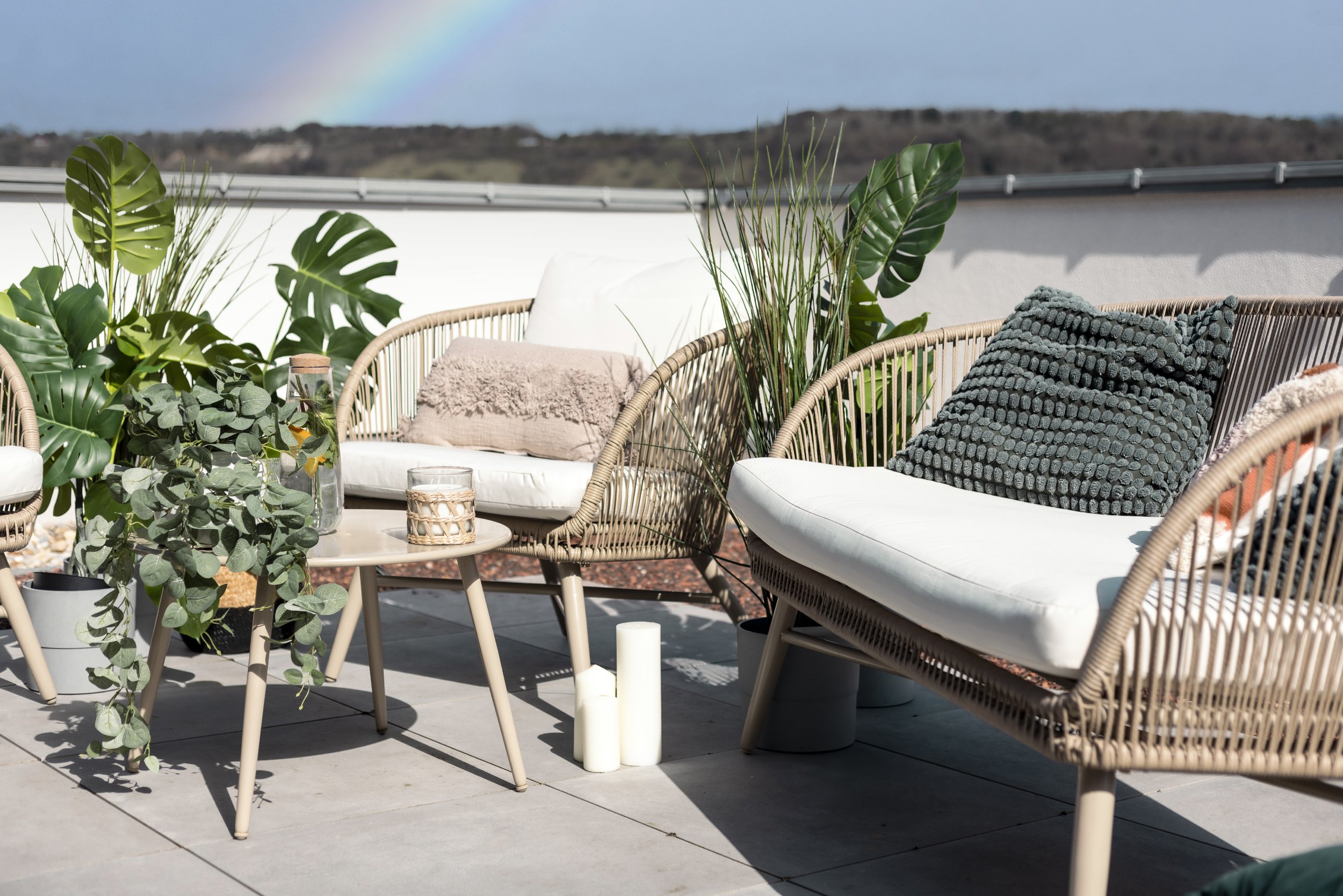 Outdoor patio with wicker furniture, cushions, potted plants, and a rainbow in the sky.