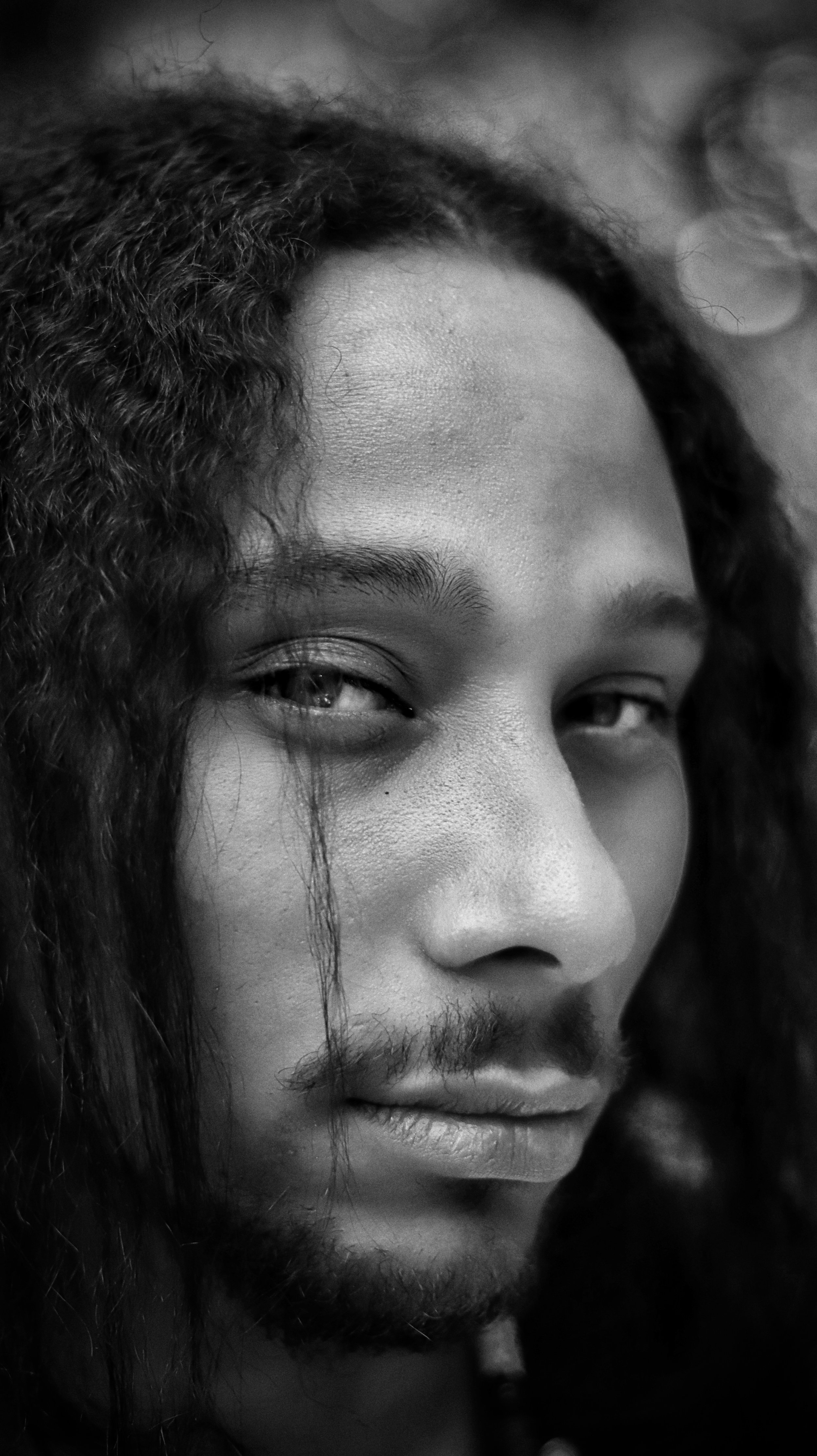 Close-up black and white portrait of a person with long curly hair and a light beard, gazing intently at the camera.