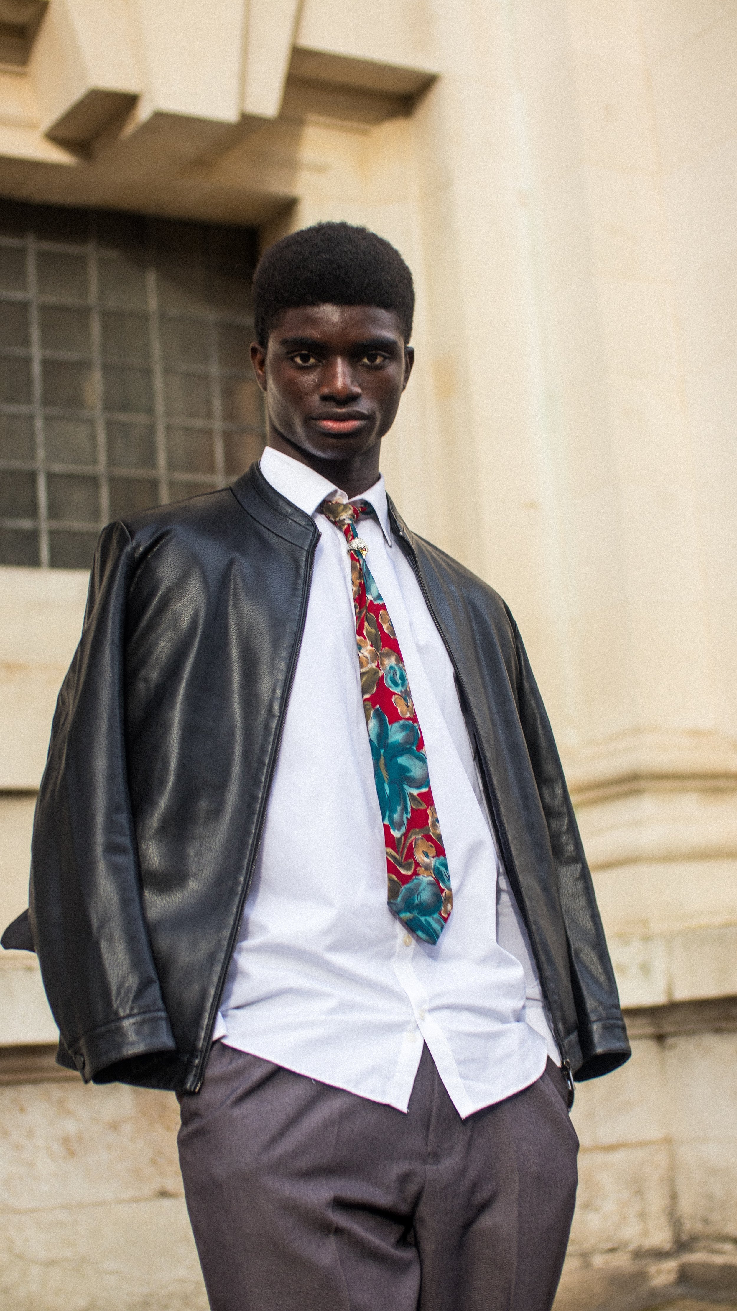 Man wearing a black leather jacket, white shirt, and colorful floral tie standing in front of a beige building.