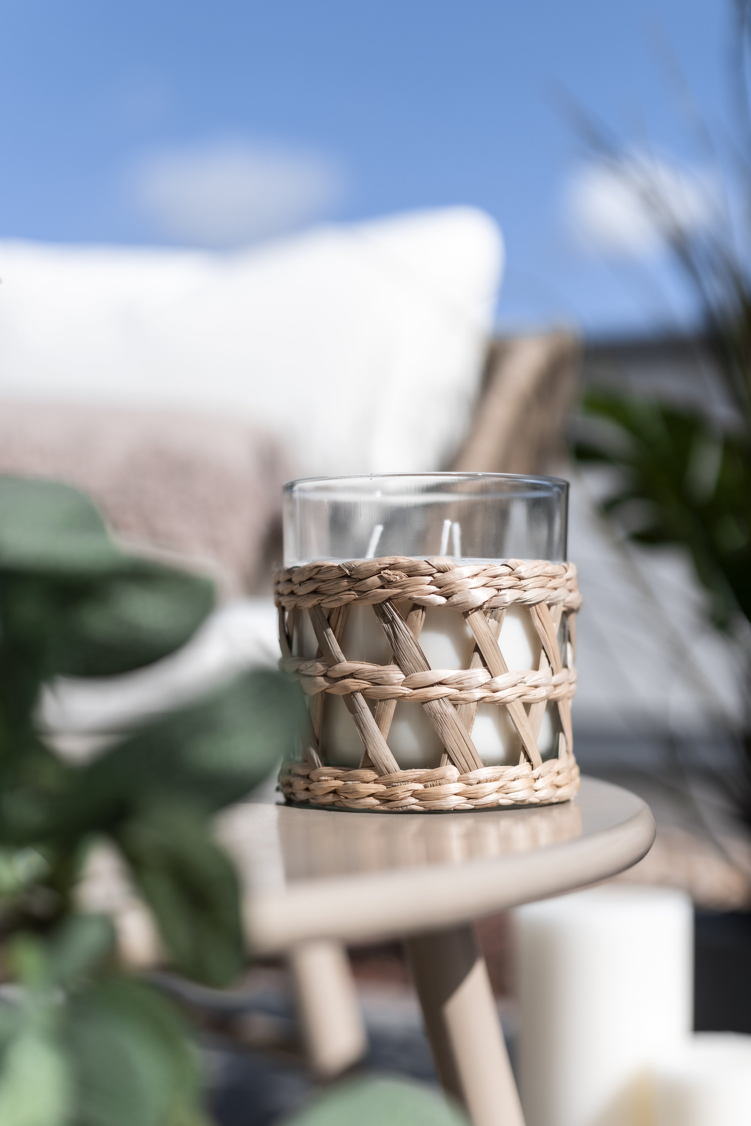 Close-up of candle in wicker holder on outdoor table with blurred green leaves, blue sky, and white pillow in the background.