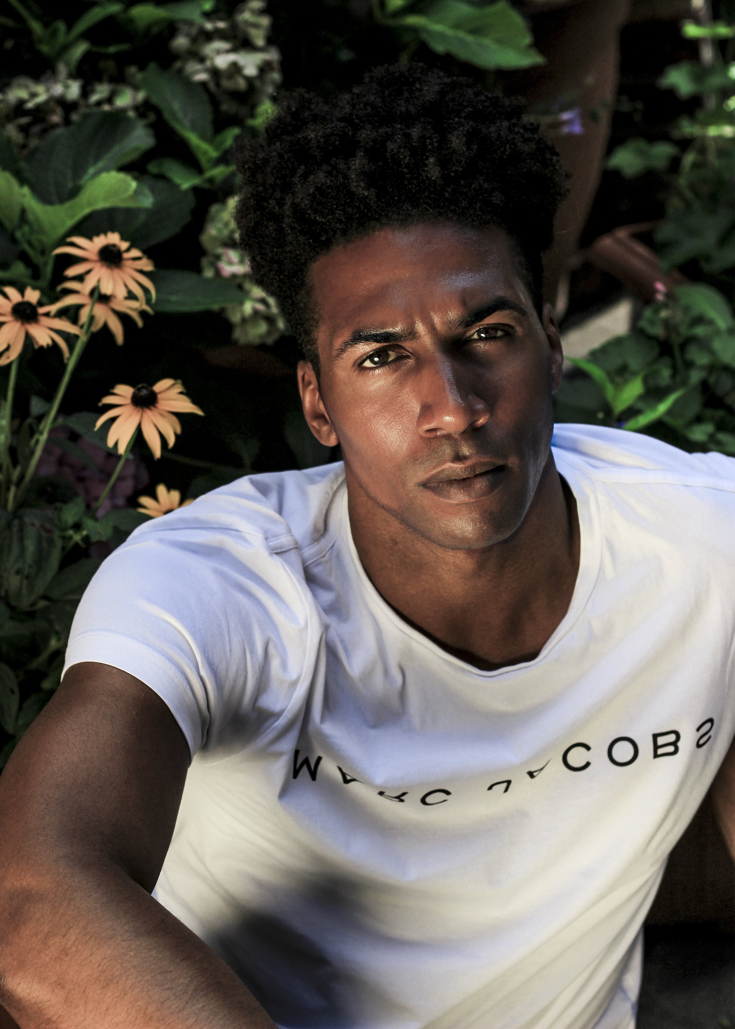 Person in white T-shirt sitting in garden with yellow flowers and green foliage.