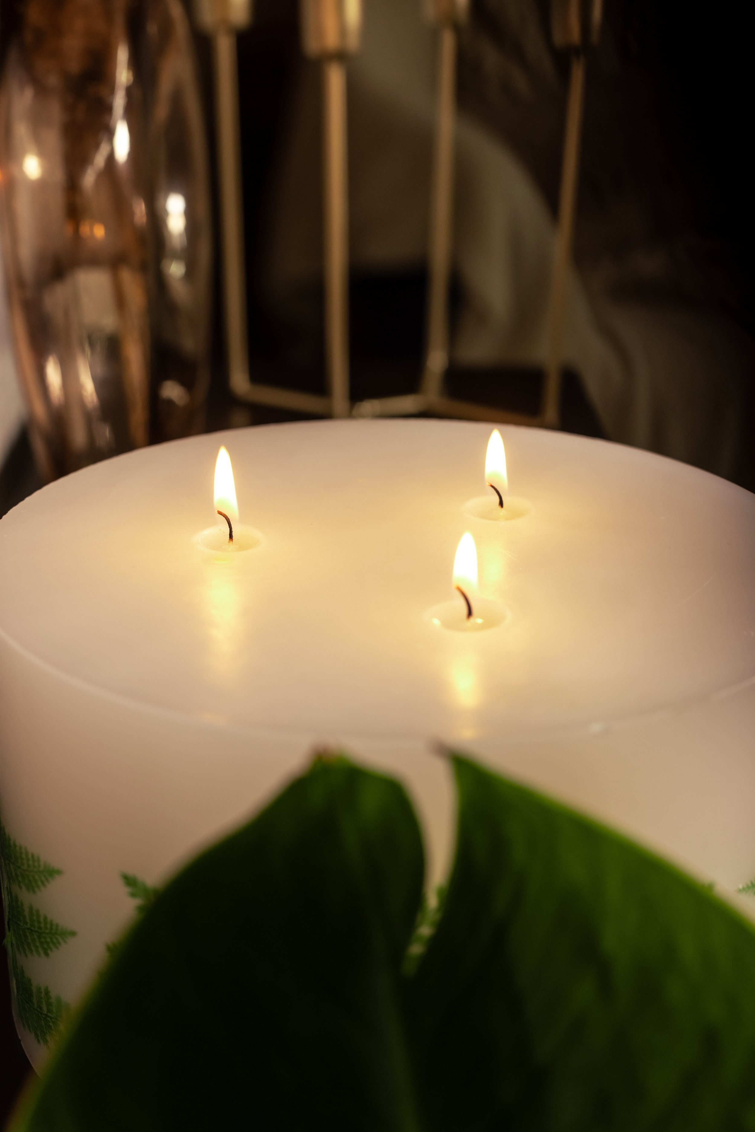 A lit triple-wick candle with fern leaf design, surrounded by blurred background elements and a green plant leaf in the foreground.