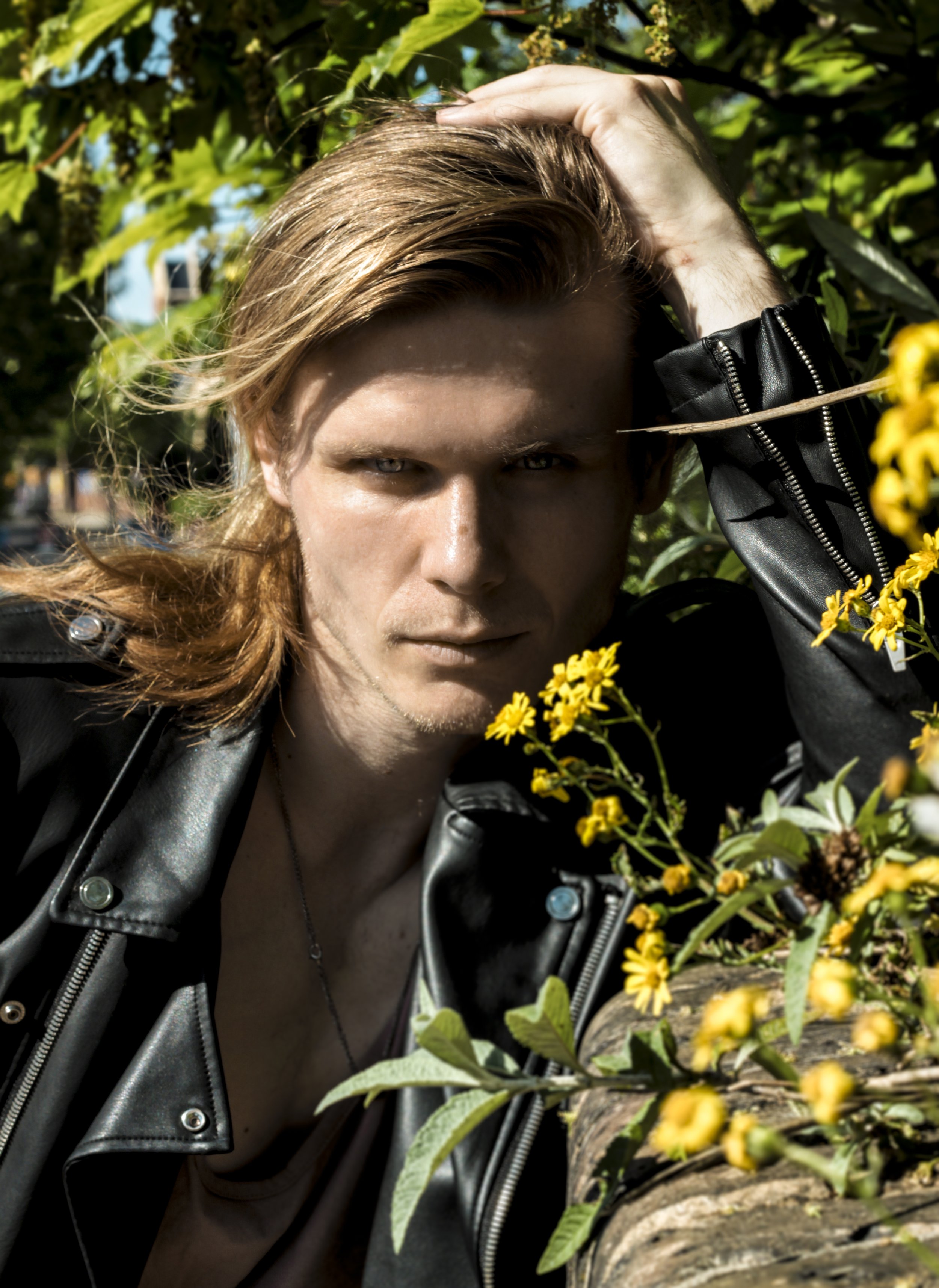 Person with long hair wearing a black leather jacket standing among yellow flowers and green leaves outdoors.