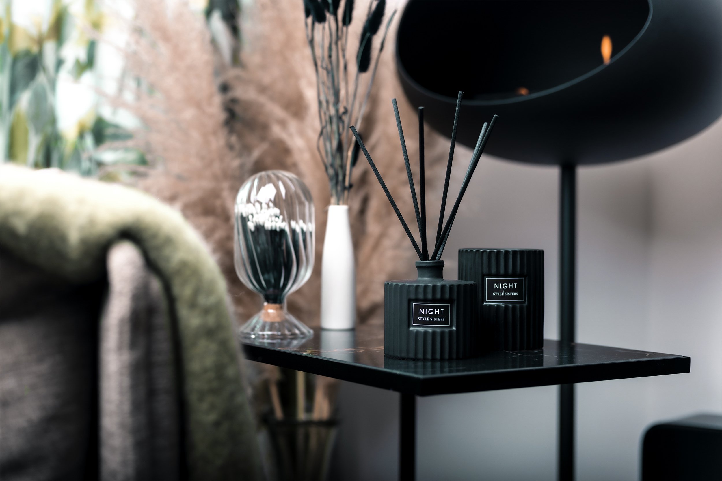 Modern interior with scented reed diffusers and hourglass on a table, surrounded by decorative plants and a lamp.