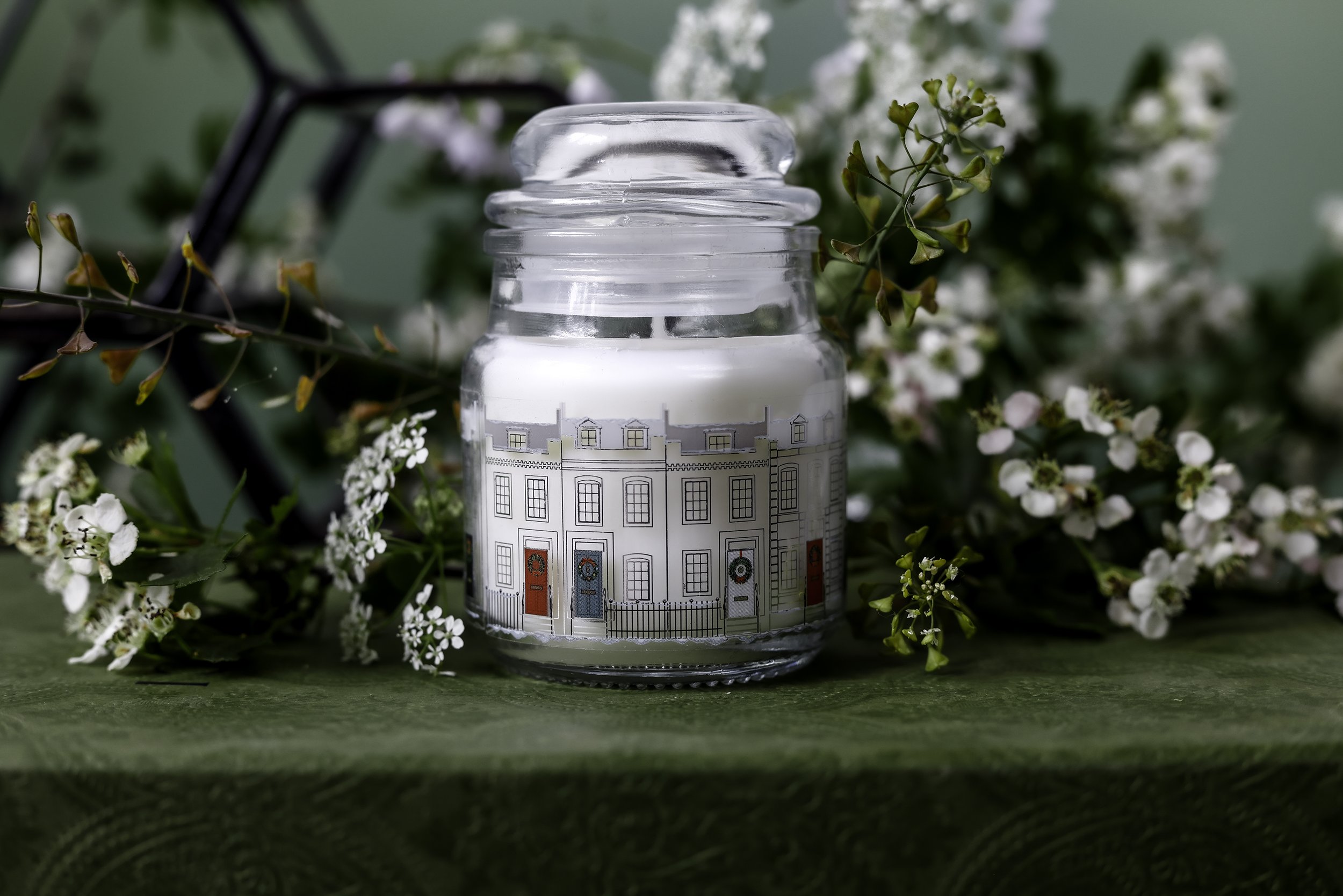 A decorative glass jar candle with a painted design of houses, surrounded by small white flowers and greenery.