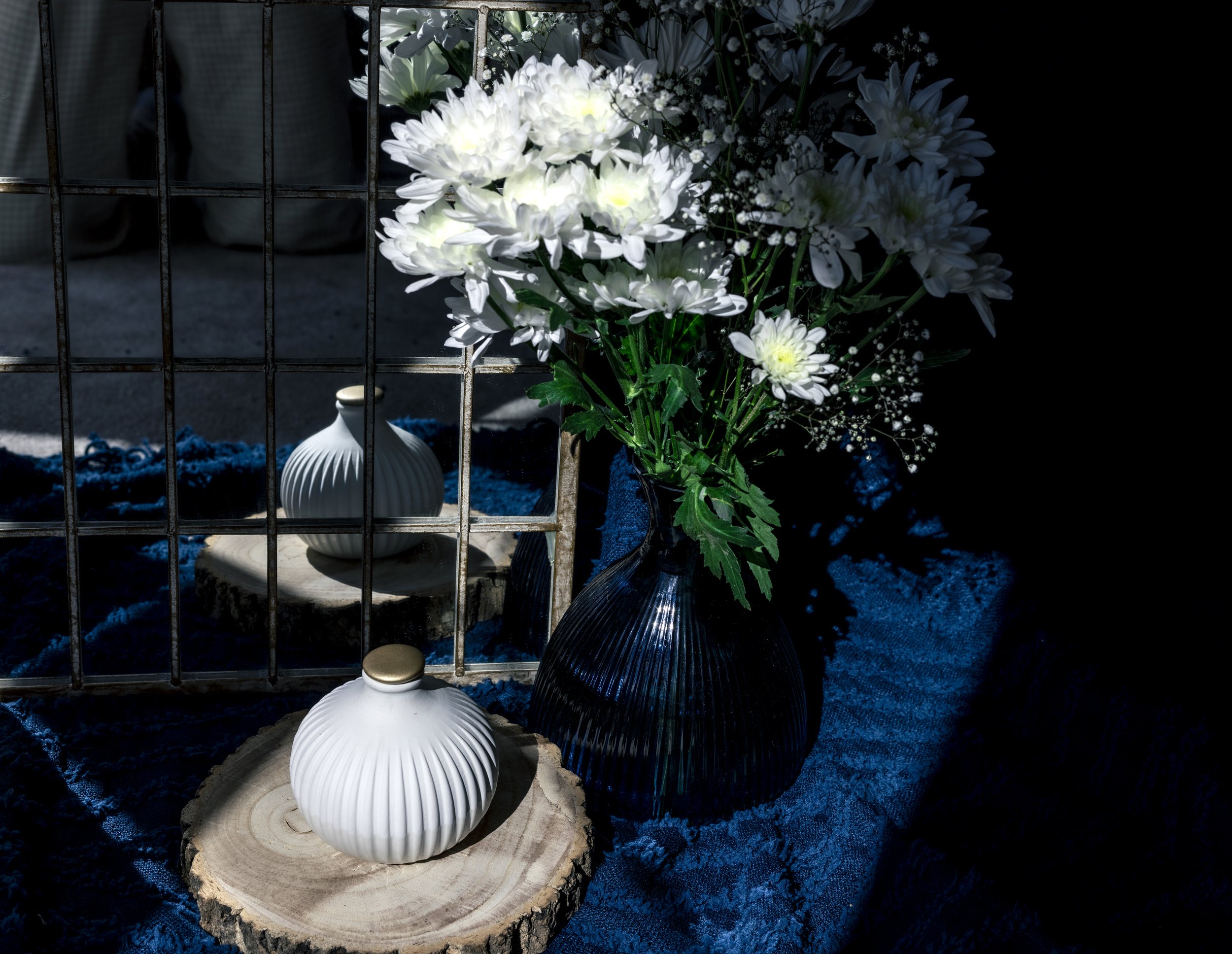 A decorative setup with a white ribbed vase on a wooden slice, white flowers in a dark vase, and a grid-patterned mirror reflecting the scene, all on a dark blue textured surface.