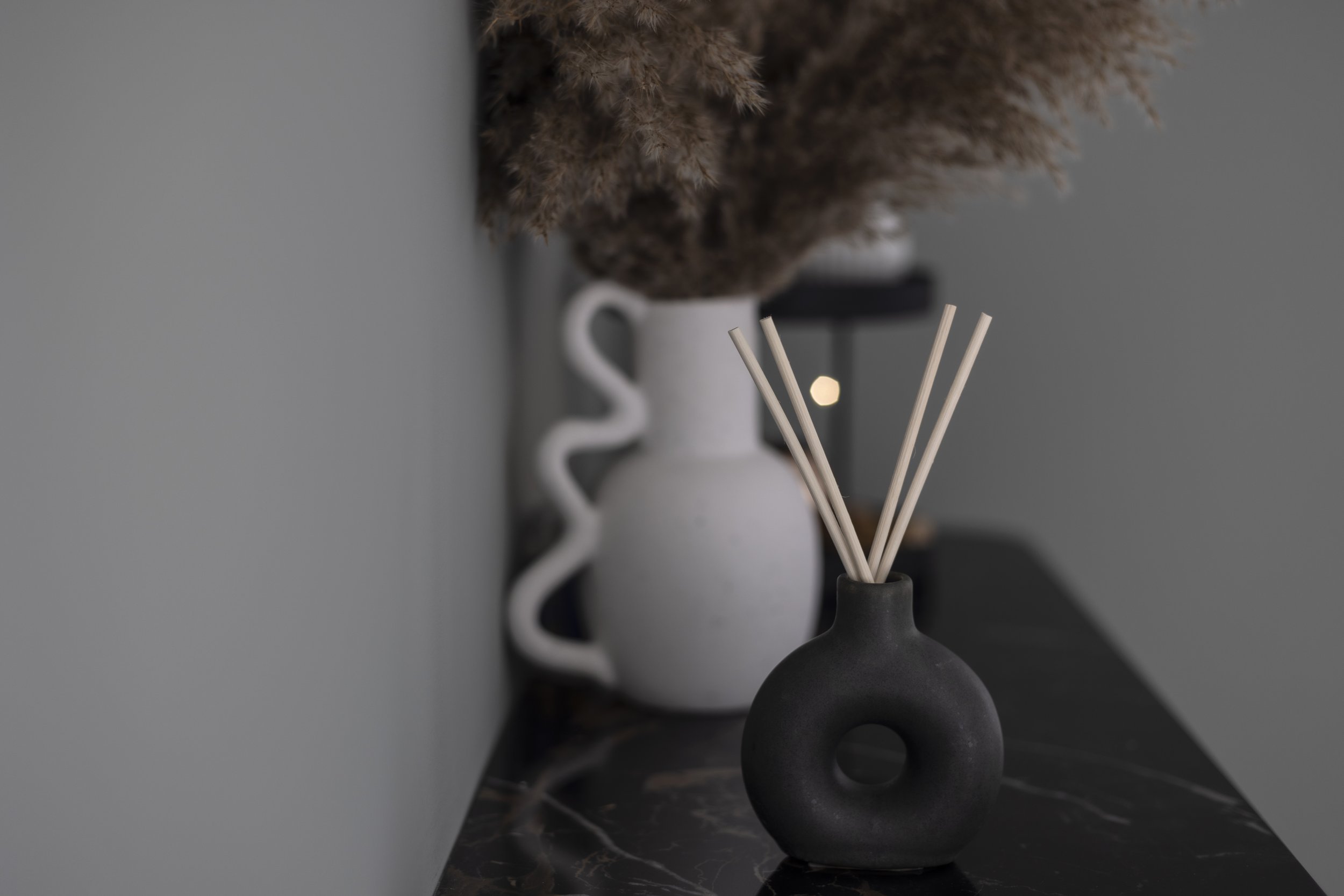 Decorative black round vase with sticks on marble surface, white vase with pampas grass in background