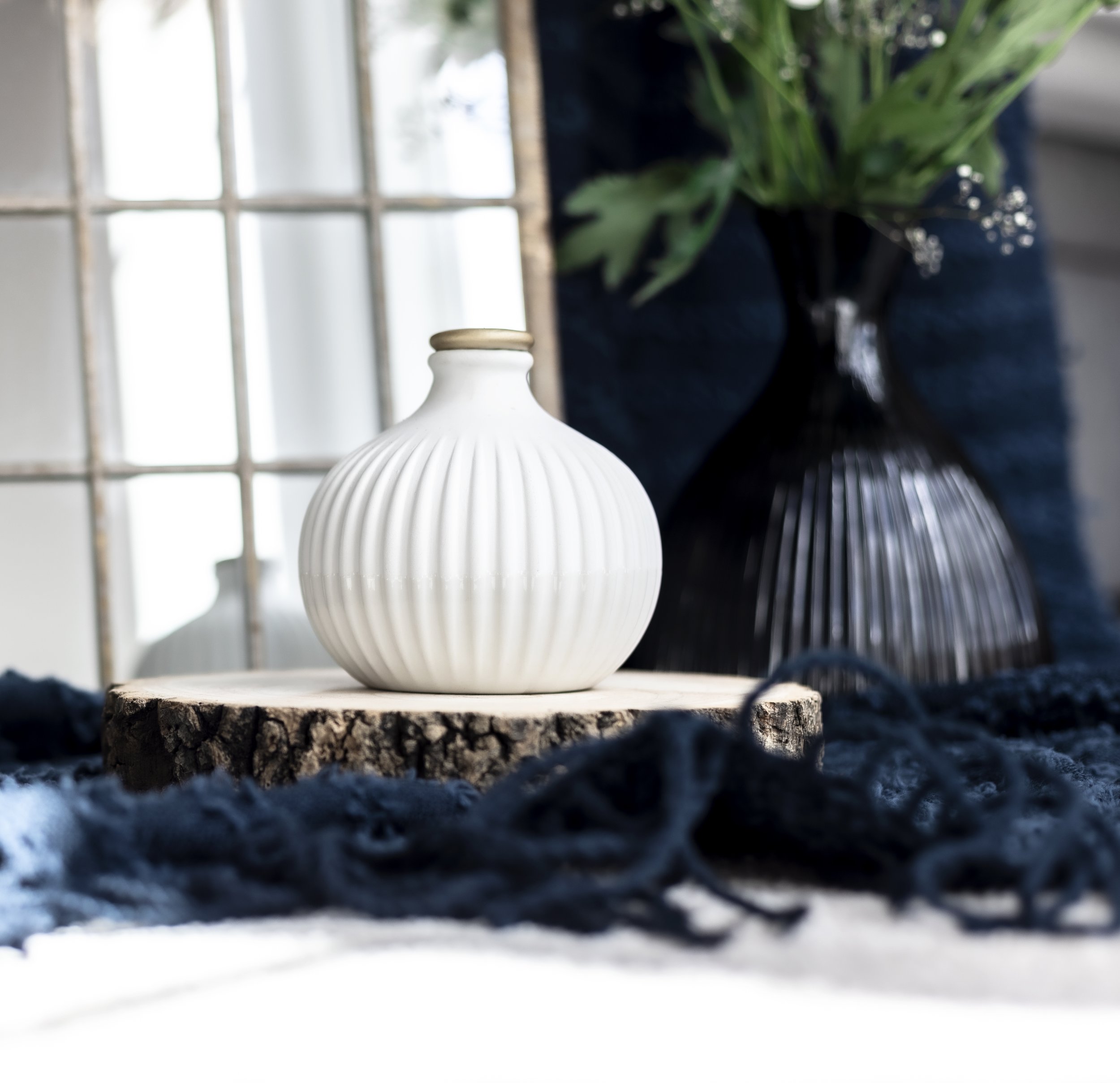 White decorative vase on wooden surface, with dark vase and green foliage in background, near window.