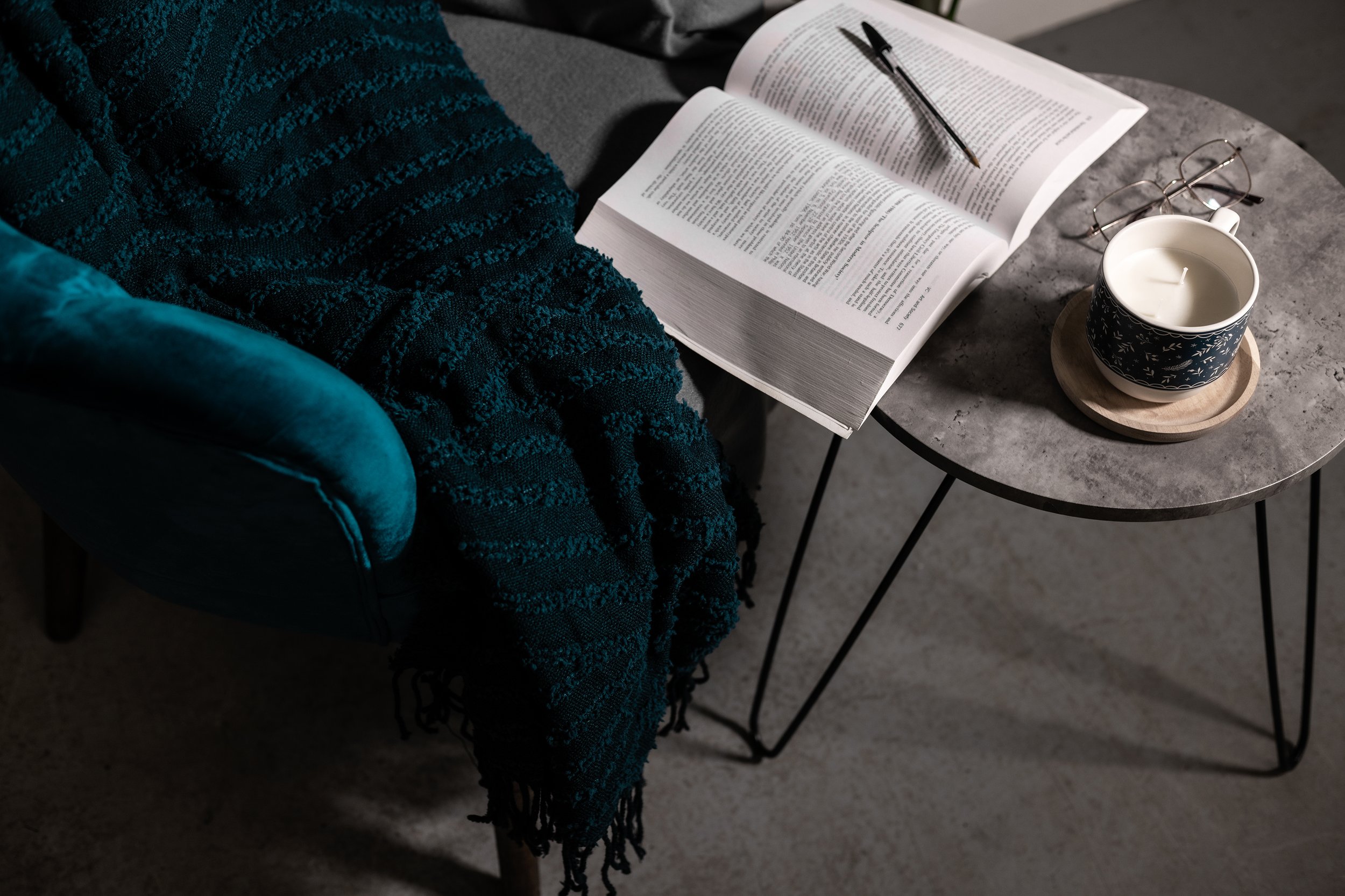 Cozy reading nook with open book, glasses, and candle on a round table. Teal blanket draped over a velvet chair.