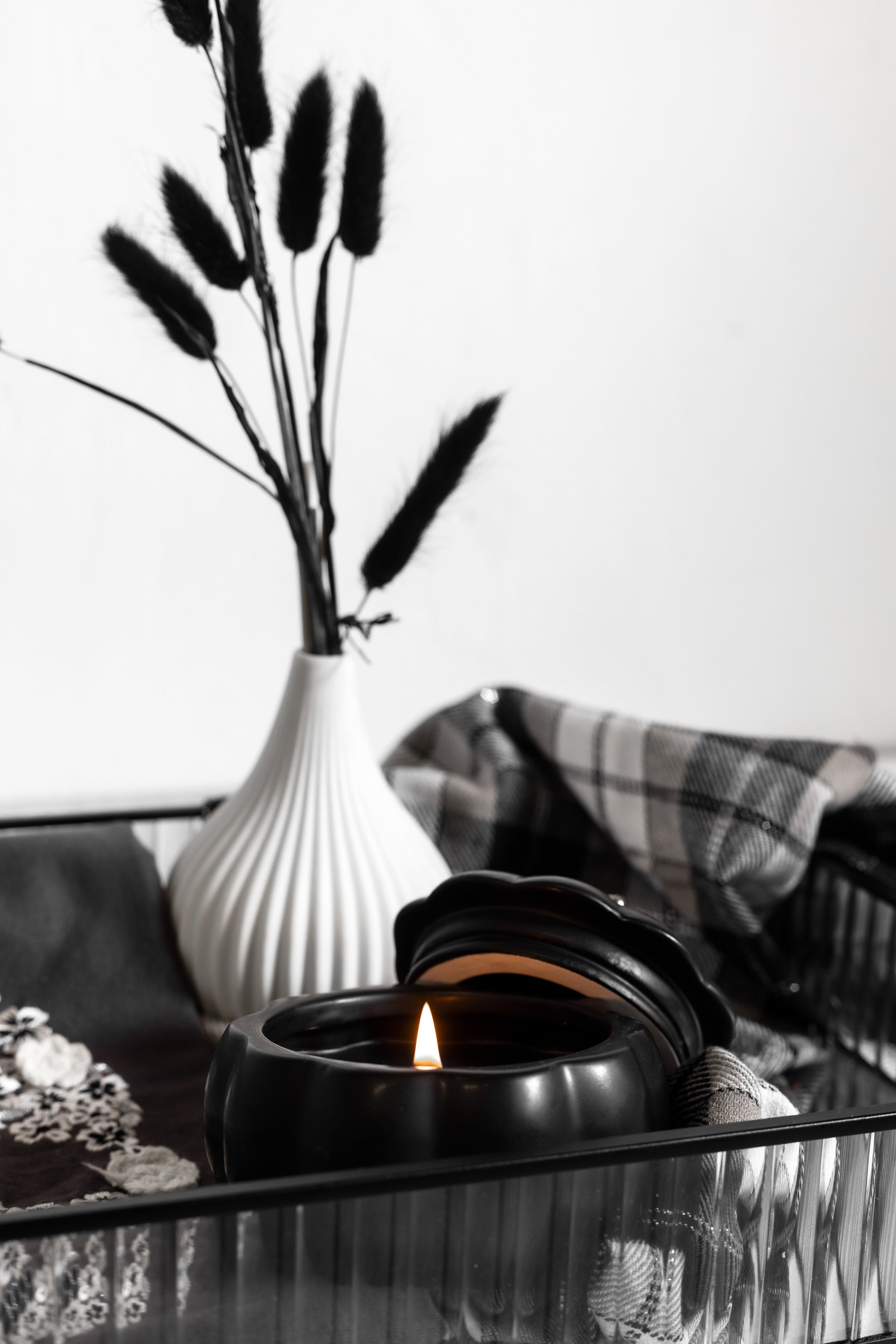 Black candle with flame beside a white vase holding dark pampas grass, placed on a tray with plaid fabric.