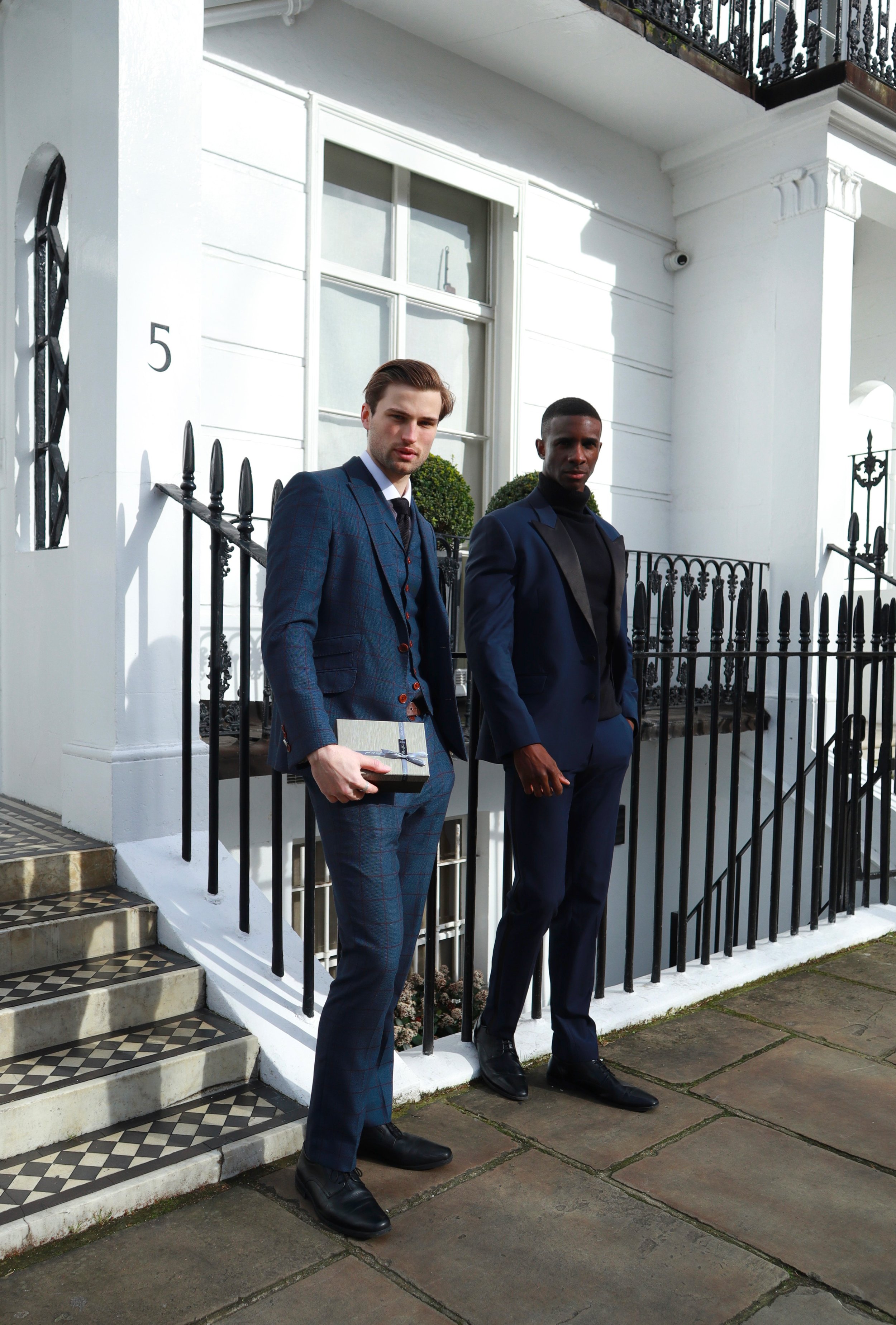 Two men in stylish suits standing outside a white townhouse with a black iron railing and tiled steps.
