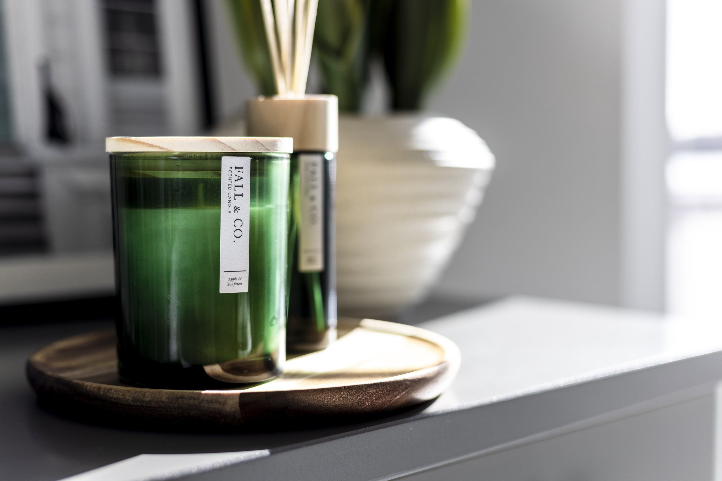 Green glass candle with "Fall & Co." label on wooden tray, next to a diffuser and white vase, on a gray surface in natural light.