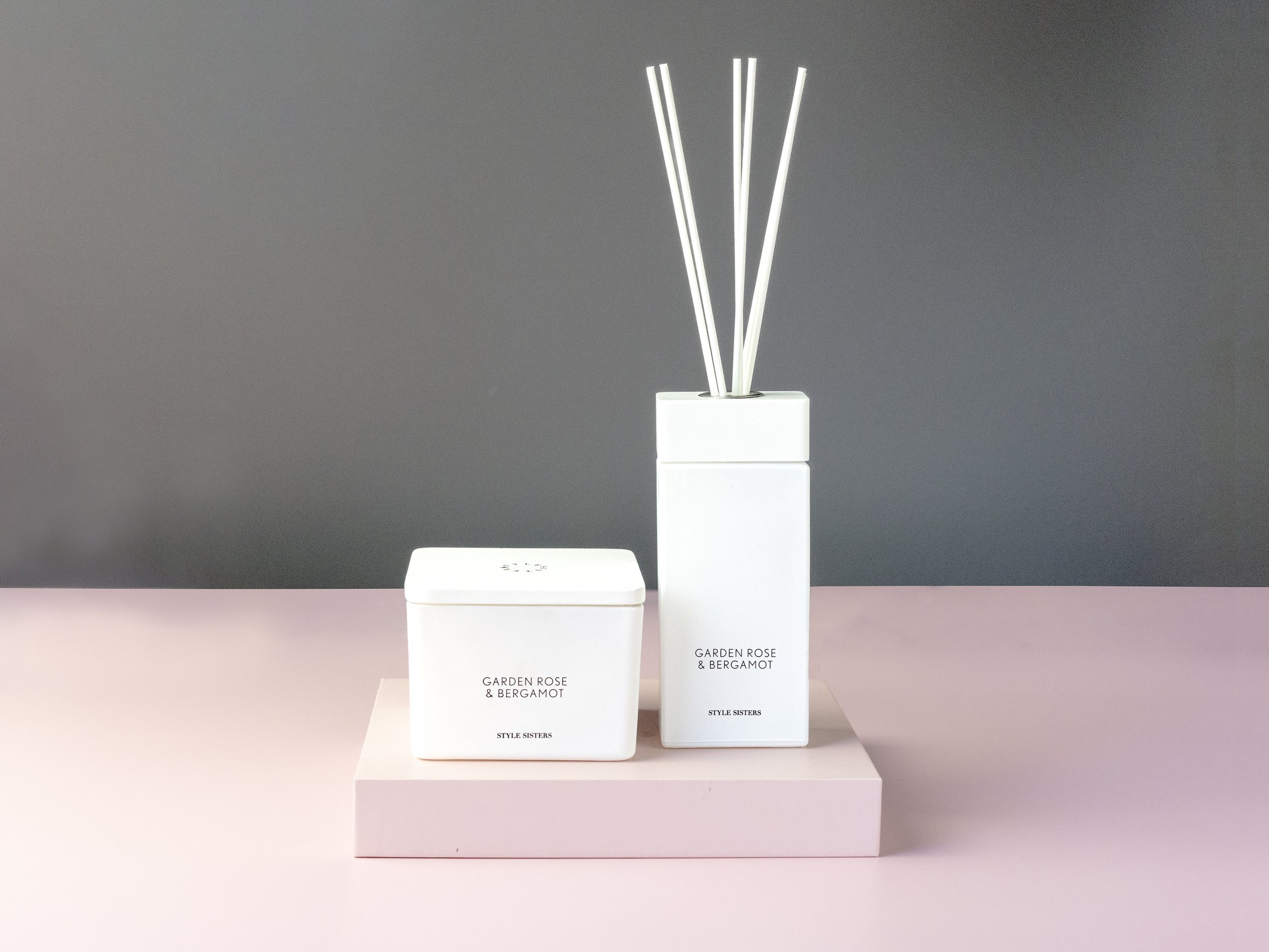 White container with "Garden Rose & Bergamot" label and white reed diffuser on a pink surface against a gray background.