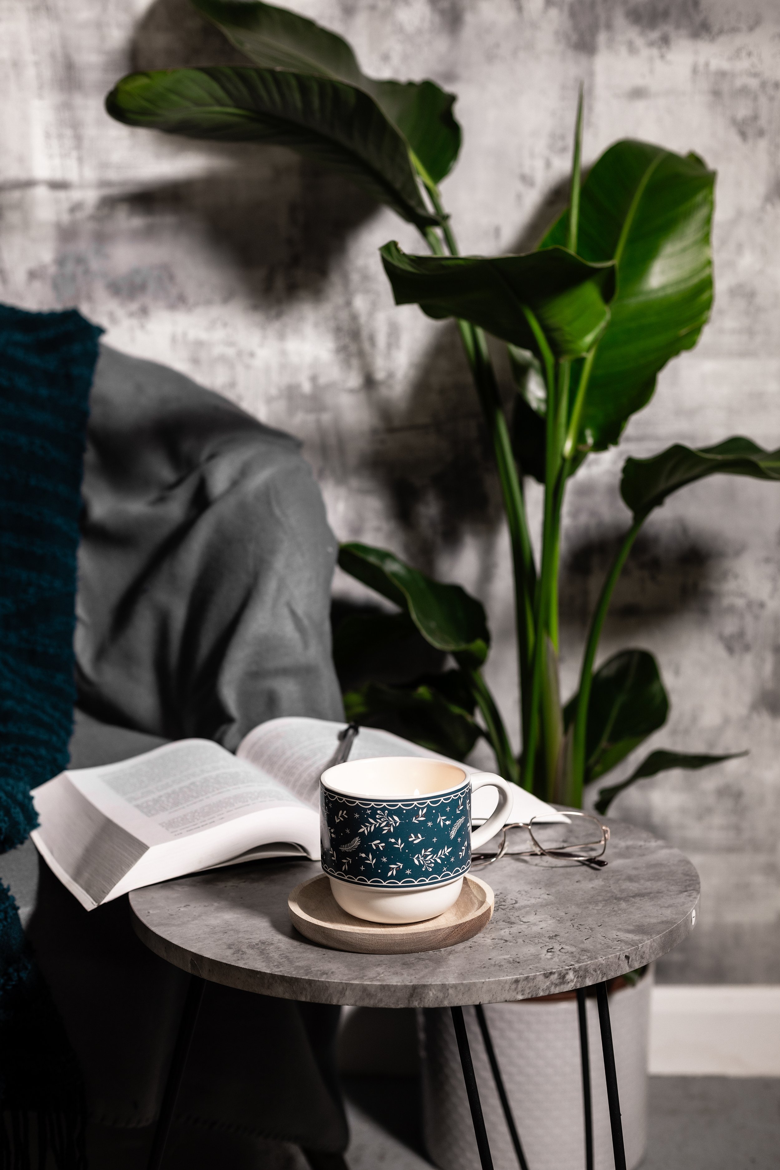 A cozy reading nook with a stone table featuring a cup on a saucer and an open book. A pair of glasses rests next to the book. Behind is a large green plant and a gray wall background.