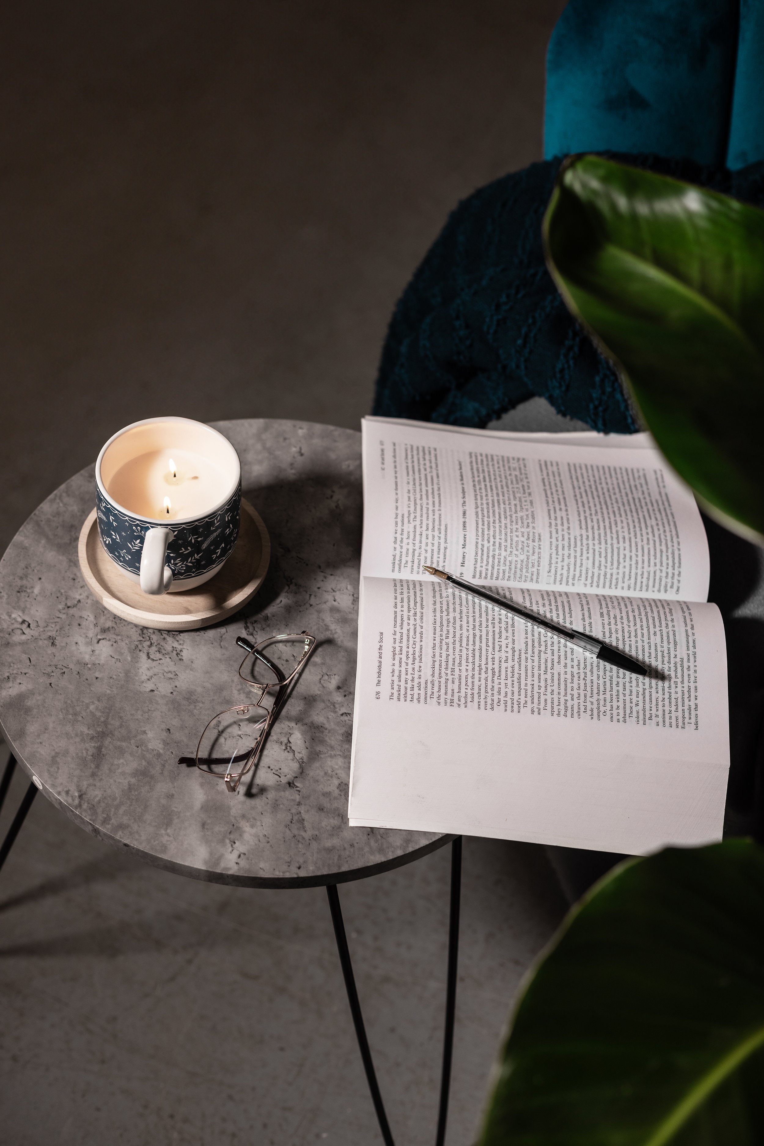 Open book with pen on a small table, next to a lit candle in a cup and reading glasses, surrounded by a teal fabric and a plant.
