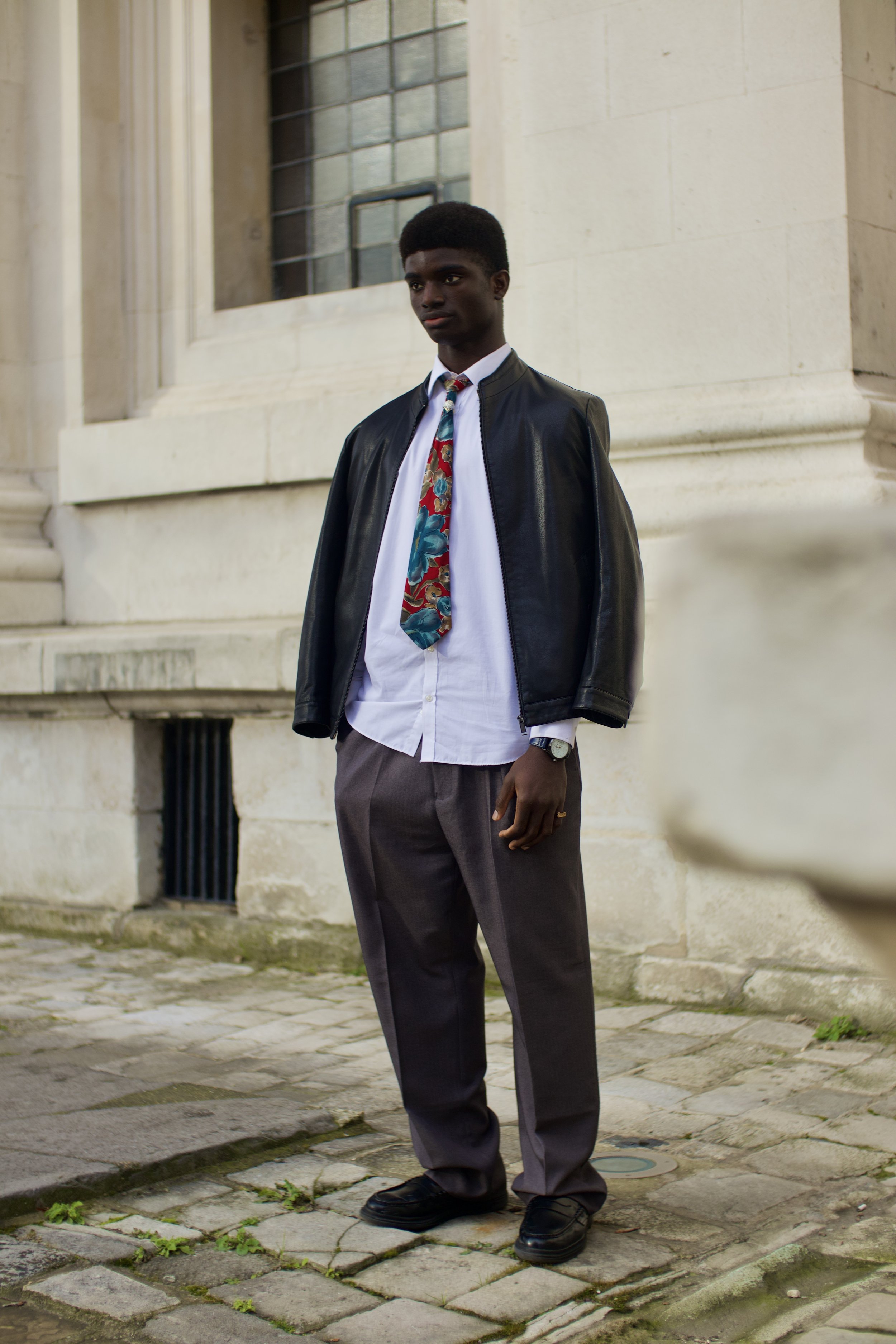 A person wearing a black leather jacket over a white shirt and colorful tie, standing outside near a stone building with large windows. They are also wearing gray pants and black shoes.