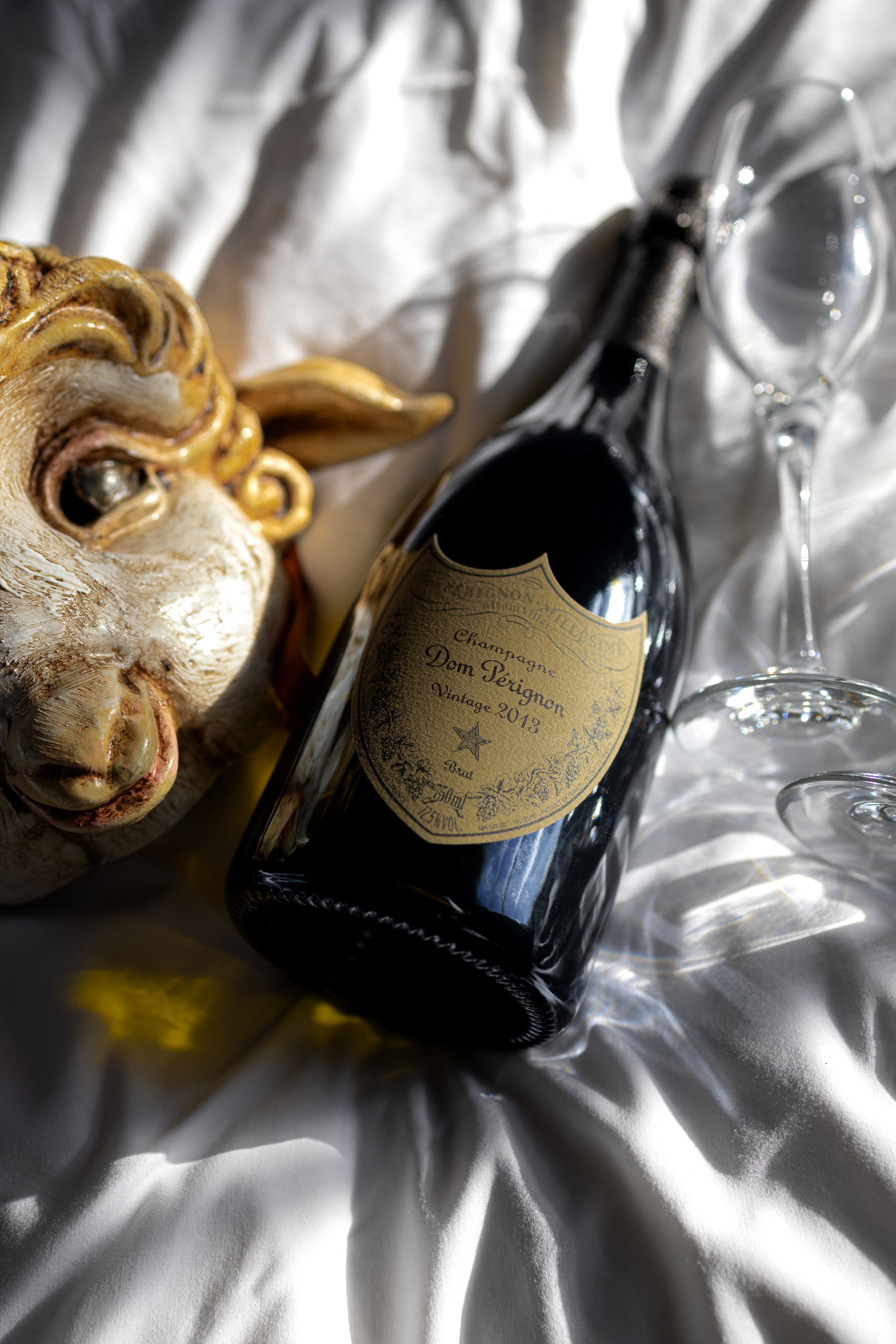 A Dom Pérignon 2013 champagne bottle on white fabric, accompanied by two empty wine glasses and a decorative mask.
