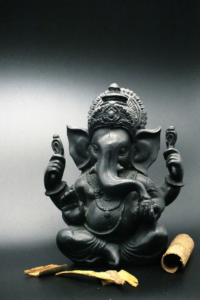 Black statue of the Hindu deity Ganesha with cinnamon sticks and bark against a dark background.