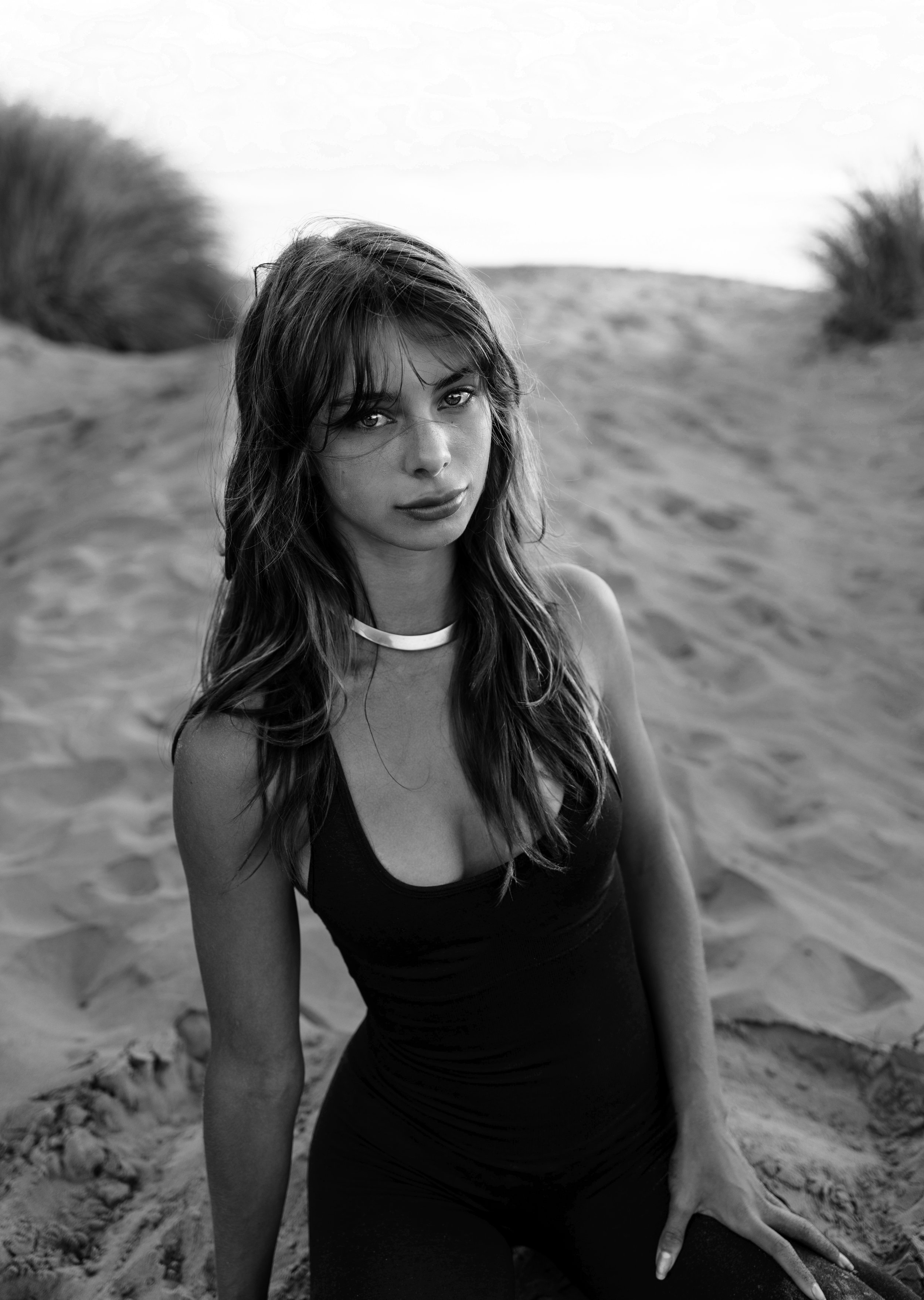 Black and white photo of a woman sitting on sand with a neutral expression, wearing a sleeveless top and necklace, with blurred vegetation in the background.