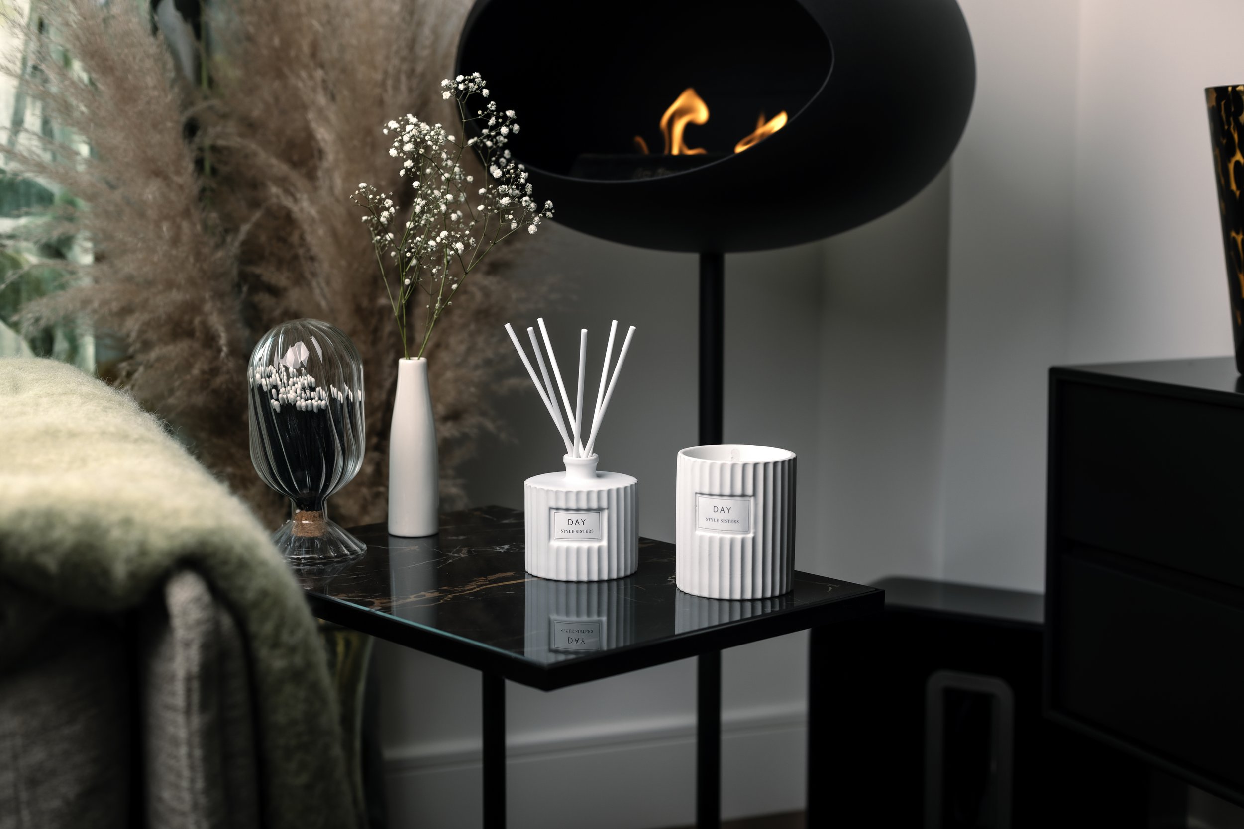 Modern interior with a black metal table holding a white diffuser, candle, and small vases with decorative foliage; a stylish chair and a small black fireplace are in the background.