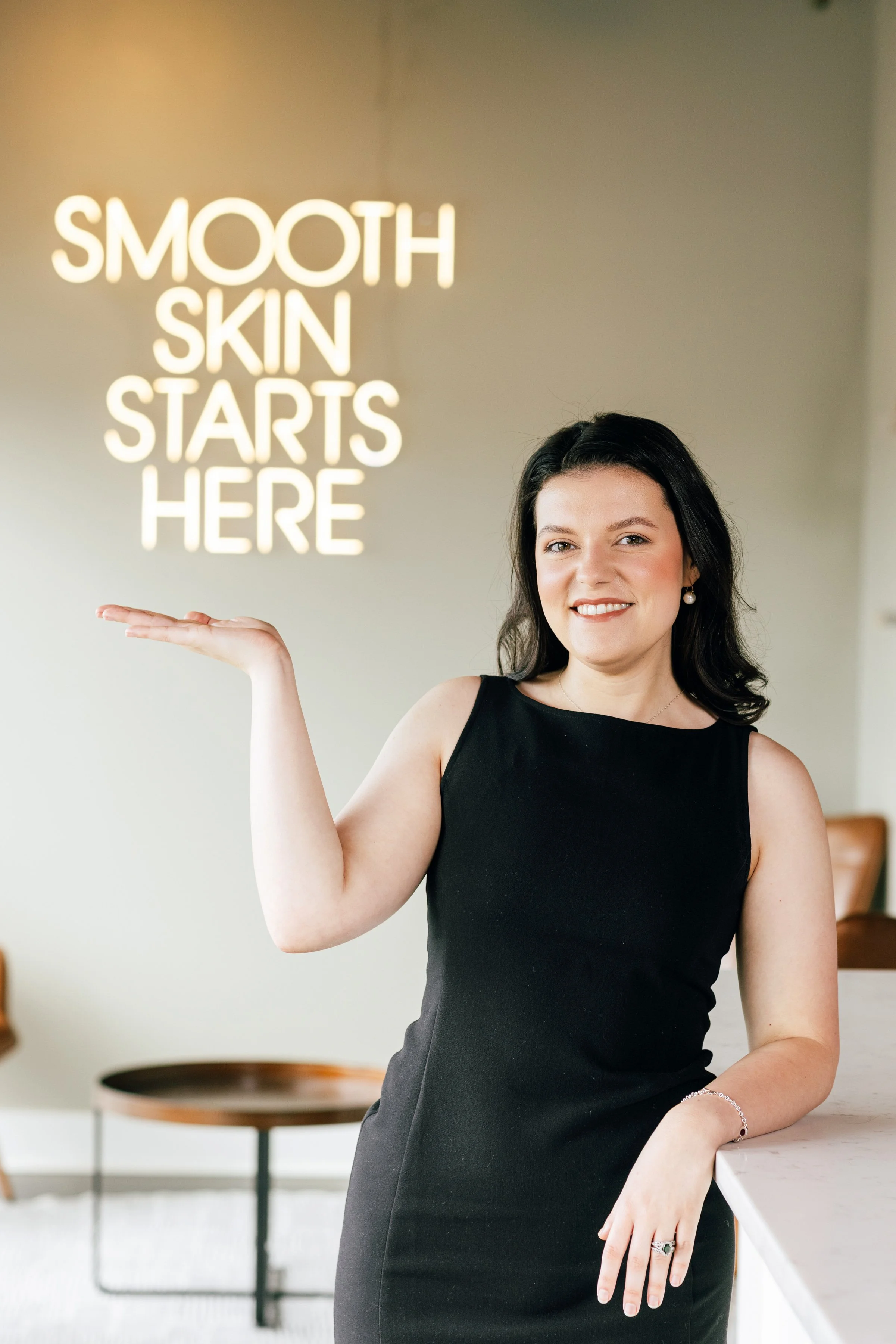A woman with dark hair and a leather jacket smiling in front of a neon sign that reads 'Smooth Skin Starts Here'.