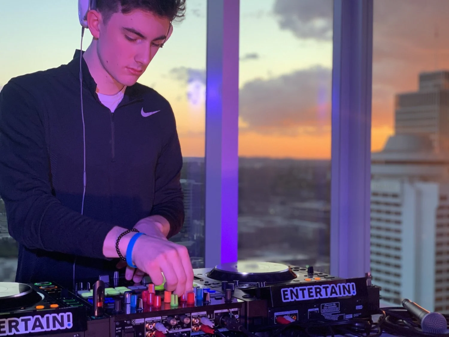A DJ is mixing music on a professional DJ controller in a high-rise with a cityscape view at sunset. He wears a black Nike jacket and headphones, with purple and orange hues in the sky visible through the windows.