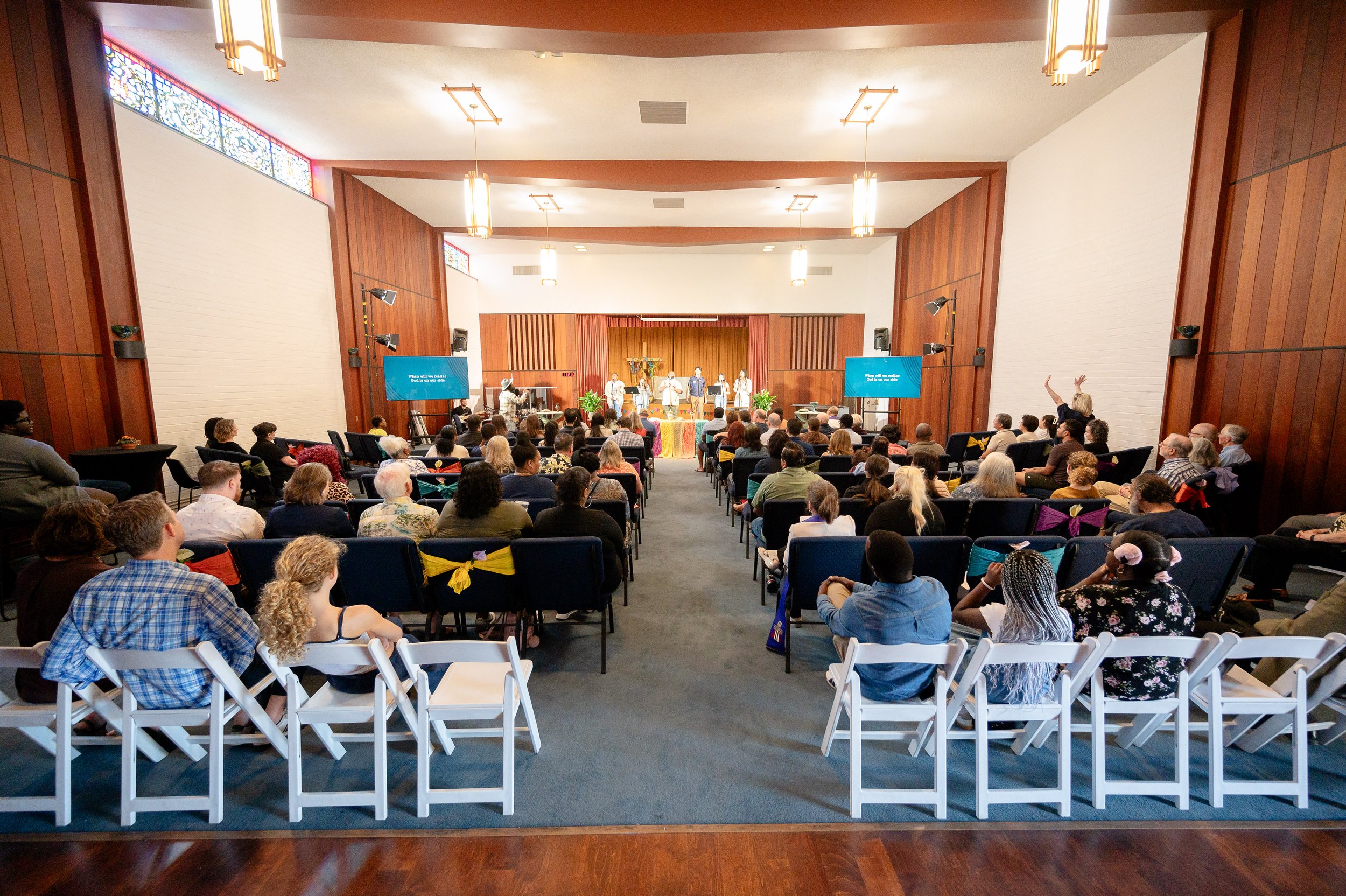 A congregation gathered in a church a service, facing a stage with people speaking. There are two large screens on either side of the stage displaying text, and the room has wooden walls and hanging lights.