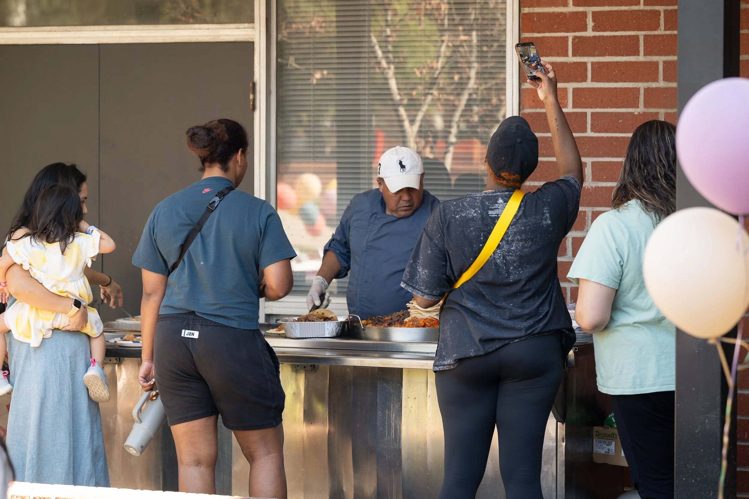 People line up at an outdoor food stand, with a server in a navy apron serving food. One woman holds her phone up to take a photo. Balloons are partially visible on the right.