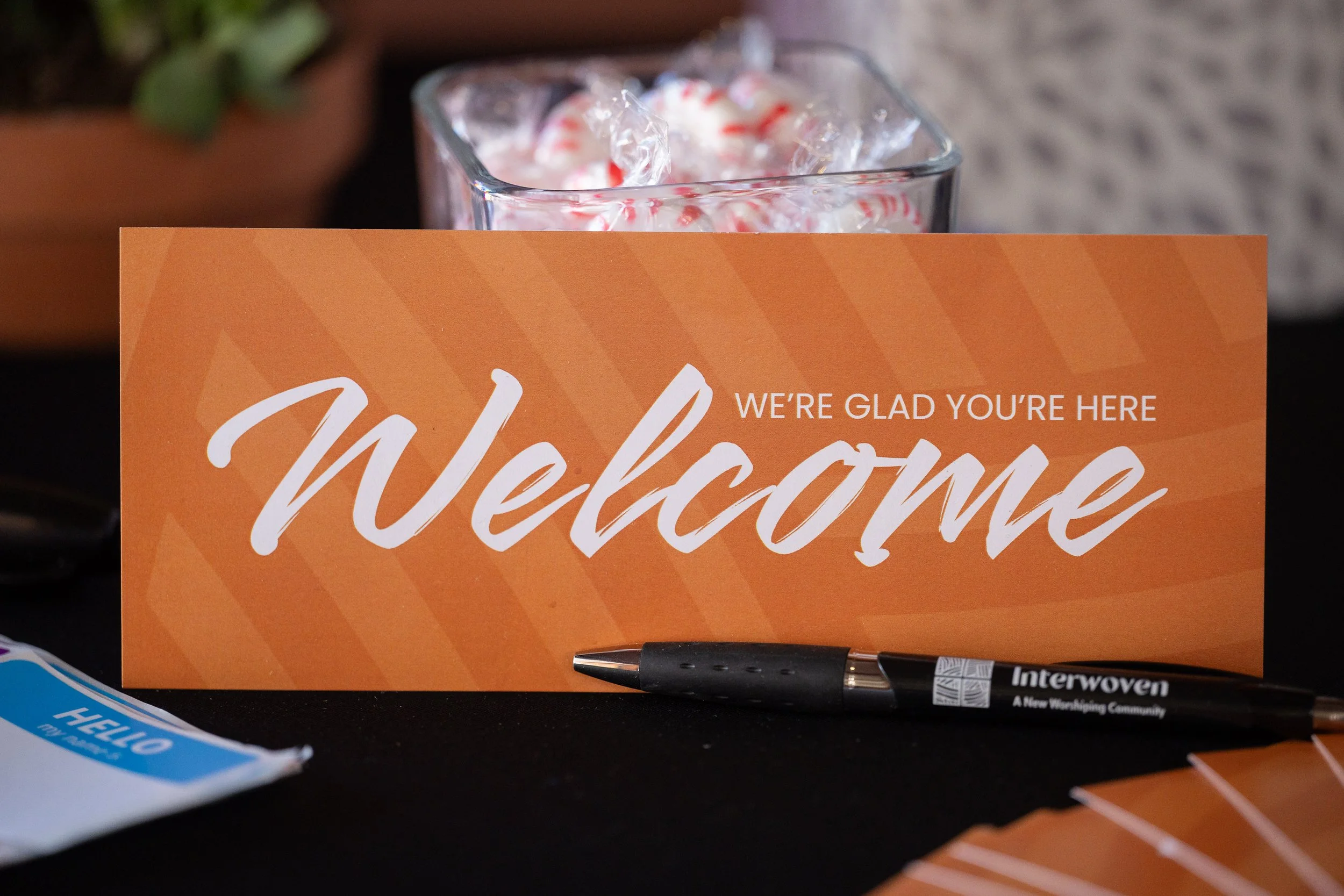 A welcome sign that says "We're glad you're here. Welcome" in white text on an orange background, with a black pen and some wrapped candies in front.