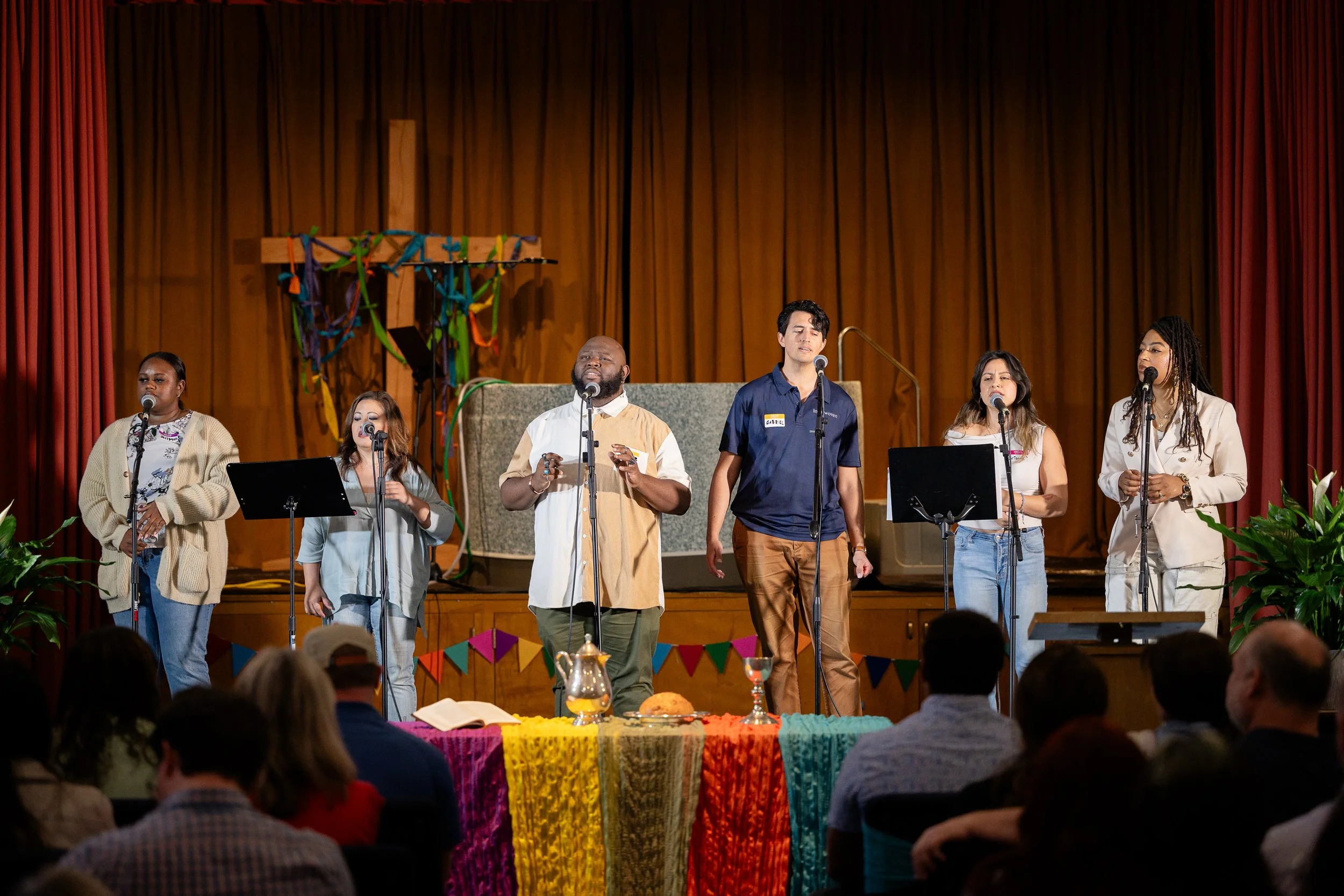 Six diverse performers singing on a stage with colorful decorations and an audience watching.