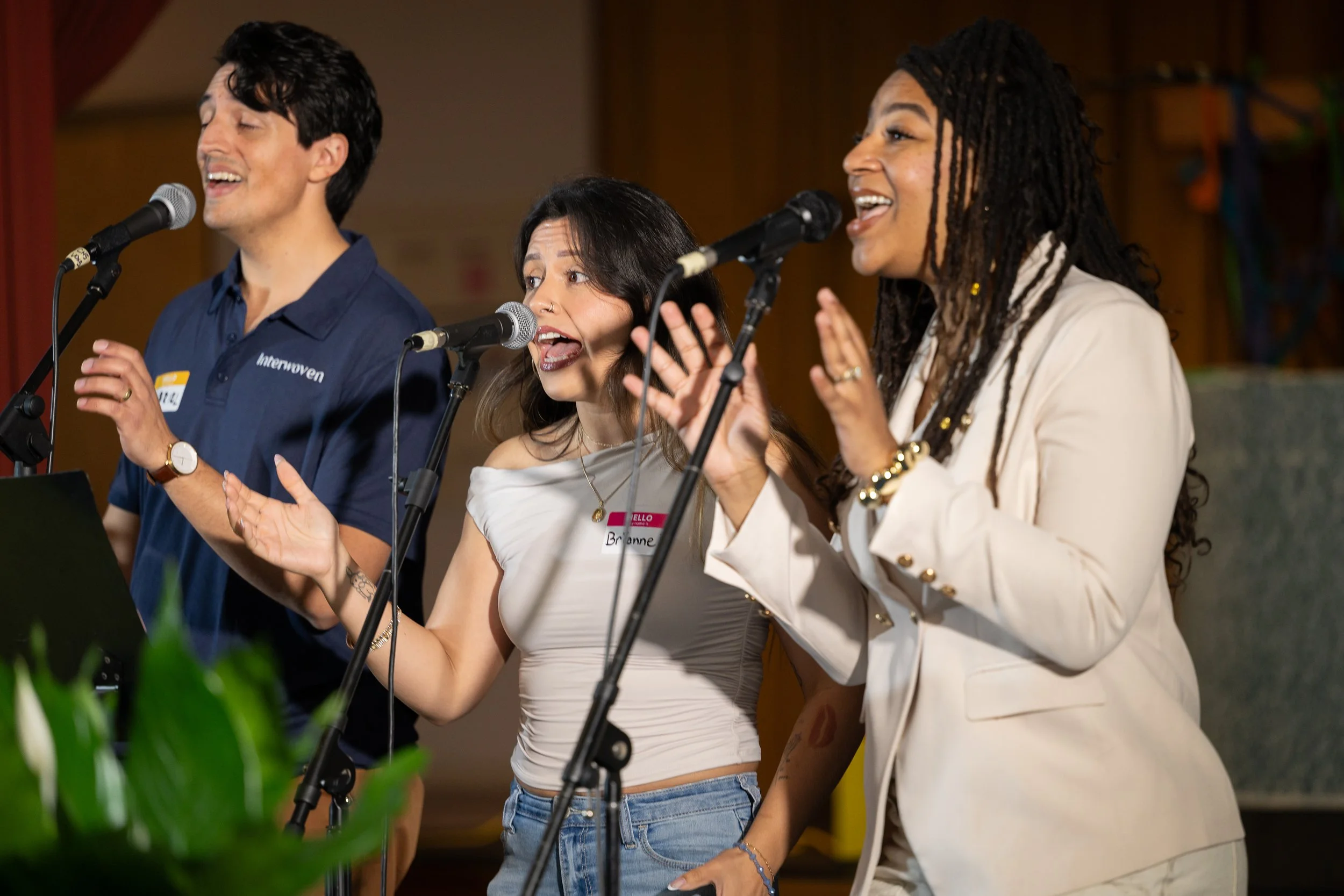 Three people singing or speaking into microphones during an event with a wooden background and some greenery in the foreground.