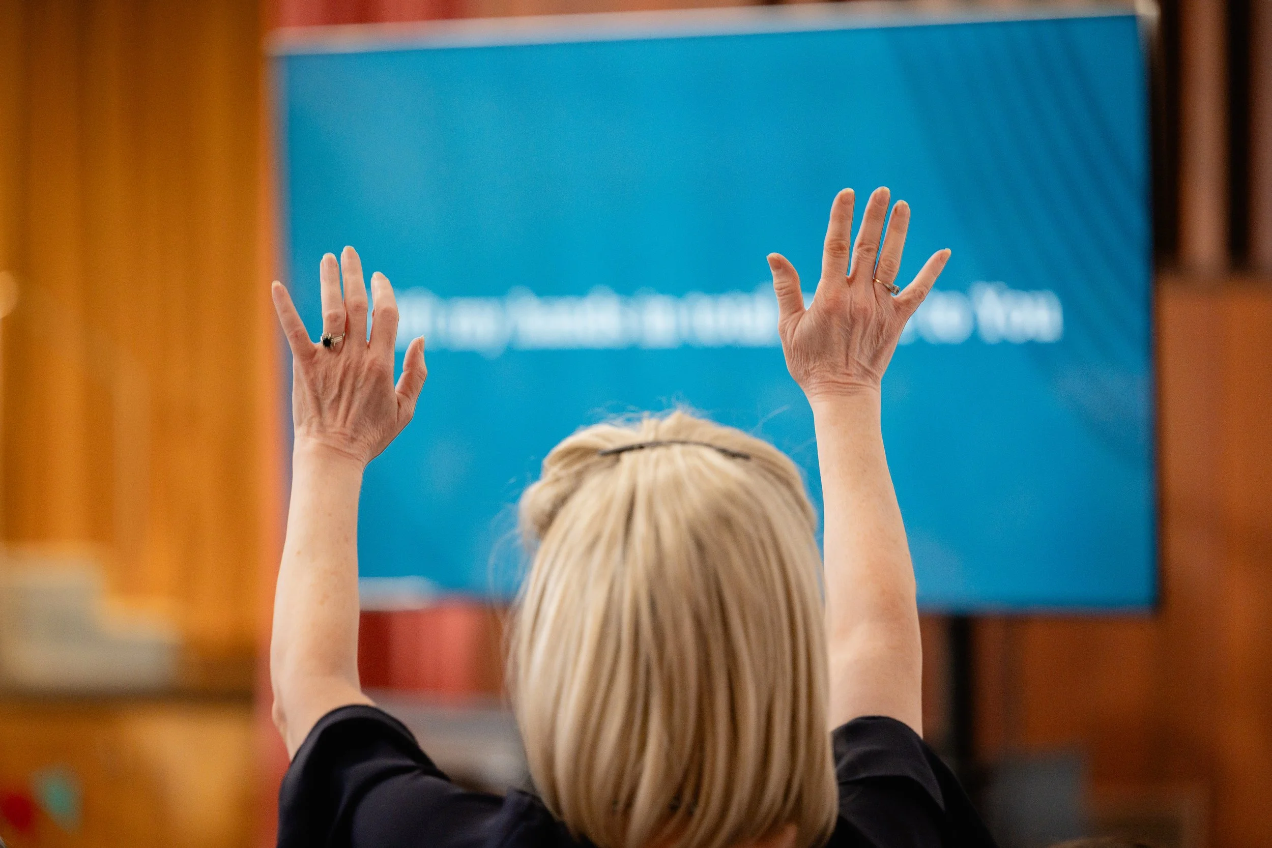 A woman with blonde hair raises her hands during a Sunday service with a large blue screen or monitor in the background.